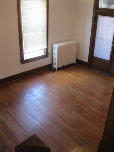 An empty room with hardwood floors, a window with blinds, a radiator, and a door with a window, all lit by natural light.