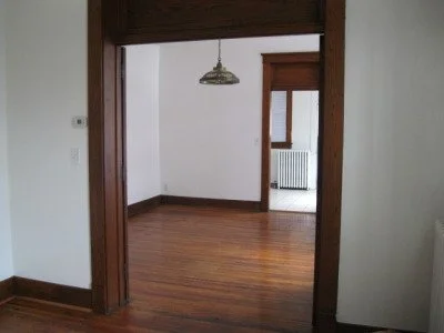 Empty room with hardwood floors and white walls, connected to another room through a wooden doorway with a hanging light fixture.