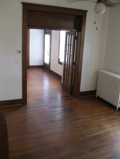 Empty room with hardwood floors, white walls, a radiator, a ceiling fan, and a doorway leading to another room with windows.