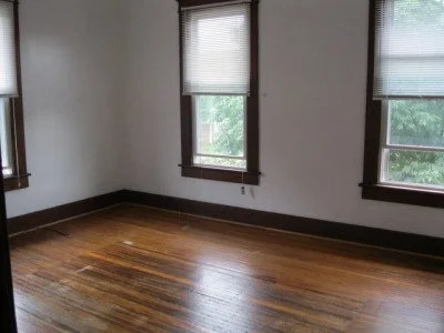 Empty room with hardwood floors and three windows with shades, overlooking trees outside.