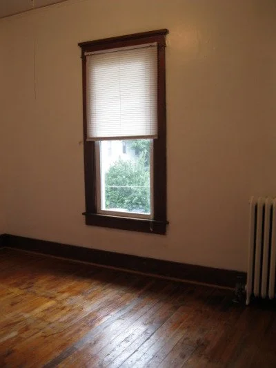 Empty room with wooden floor, single window with closed blinds, and a radiator beside the window.
