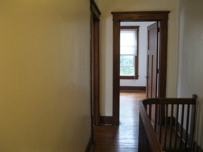 Interior view of a hallway leading to a room with a window, wooden trim, and a railing on the right side.
