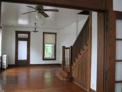 Living room with hardwood floors, wooden staircase, ceiling fan, and a window with blinds.