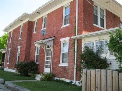 Two-story brick house with white trim, several windows, a satellite dish on the side, and bushes in the front yard.