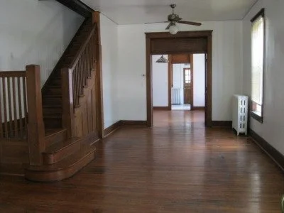 Empty living room with hardwood floors, a staircase on the left, and a doorway leading to another room in the background.