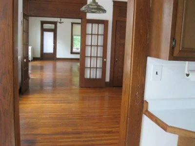 View of an empty room in a house with wood floors, a doorway with glass panes, and a kitchen area to the right with a white oven.