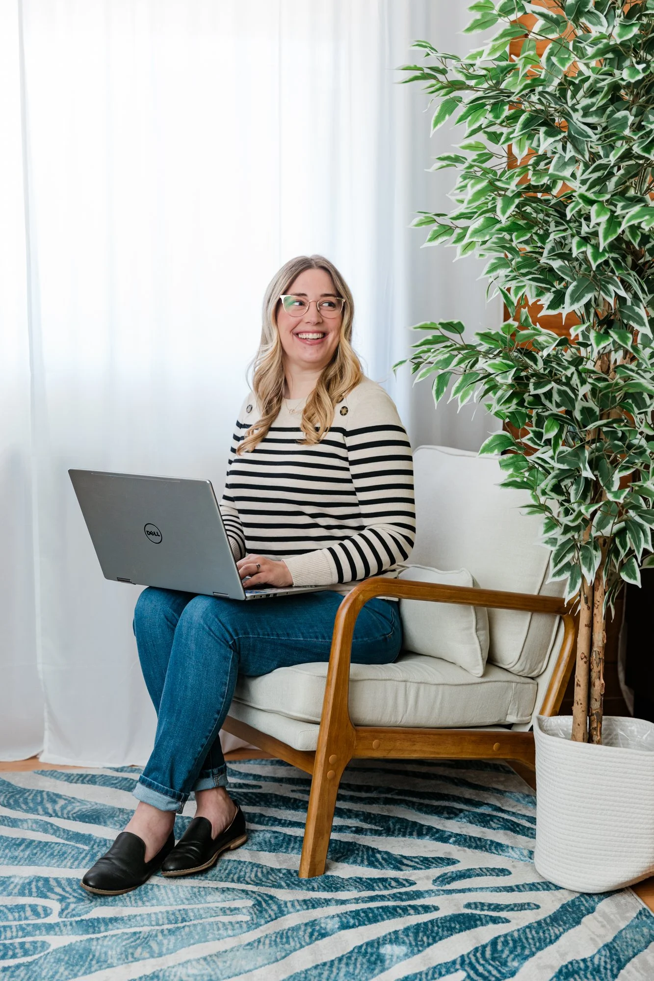 A woman smiling and sitting on a white sofa in a living room, using a laptop, with a large potted plant beside her and sheer white curtains in the background.