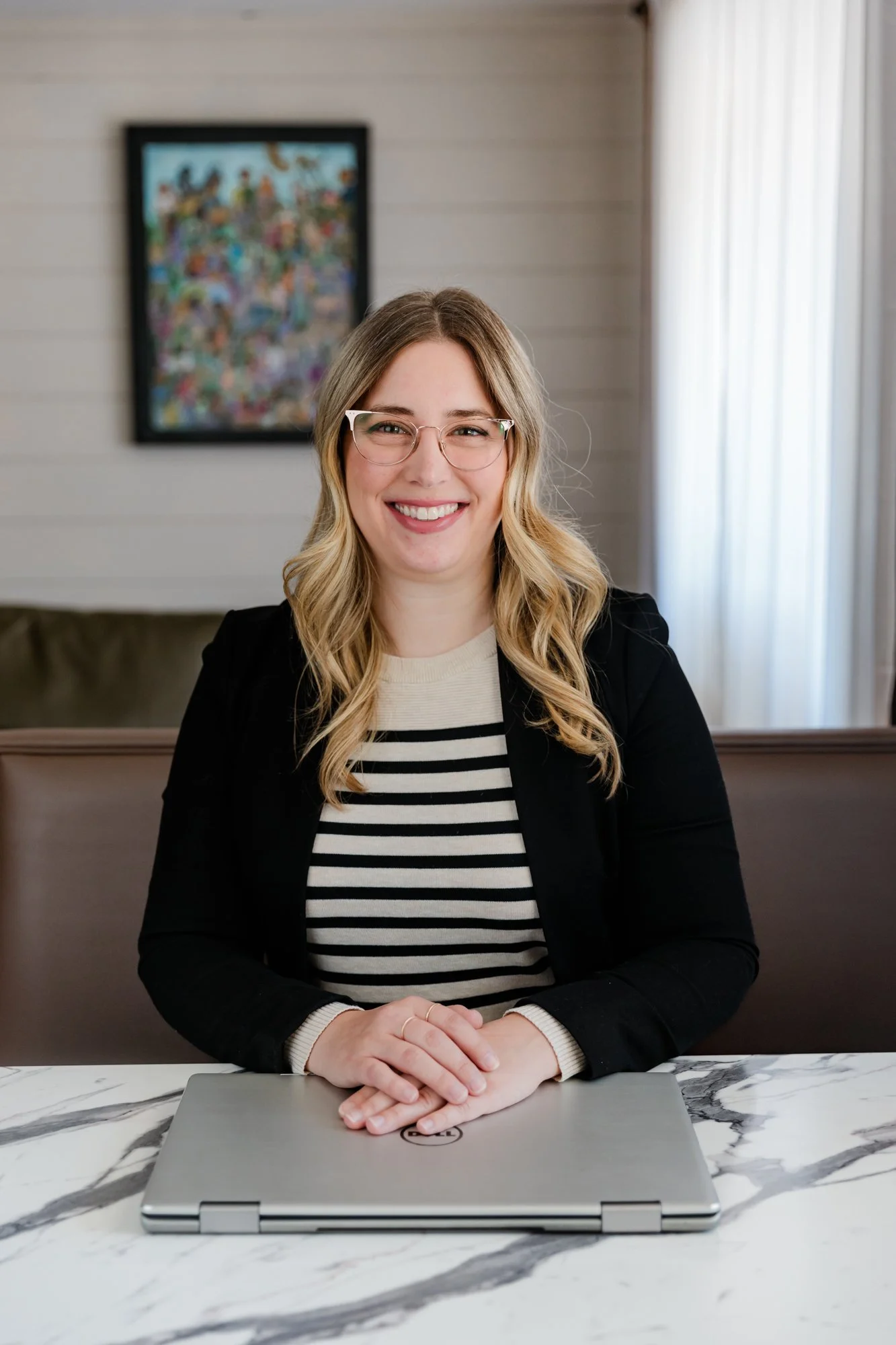 A smiling woman with glasses sitting at a marble table with a closed laptop in front of her, in a bright room with beige walls, a window with white curtains, and a framed colorful abstract picture on the wall in the background.