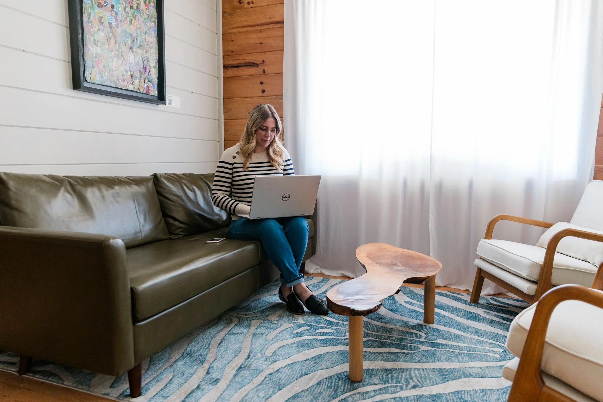 A woman with blonde hair and glasses sitting on a brown leather couch using a laptop in a cozy living room with wooden accents and modern furniture.