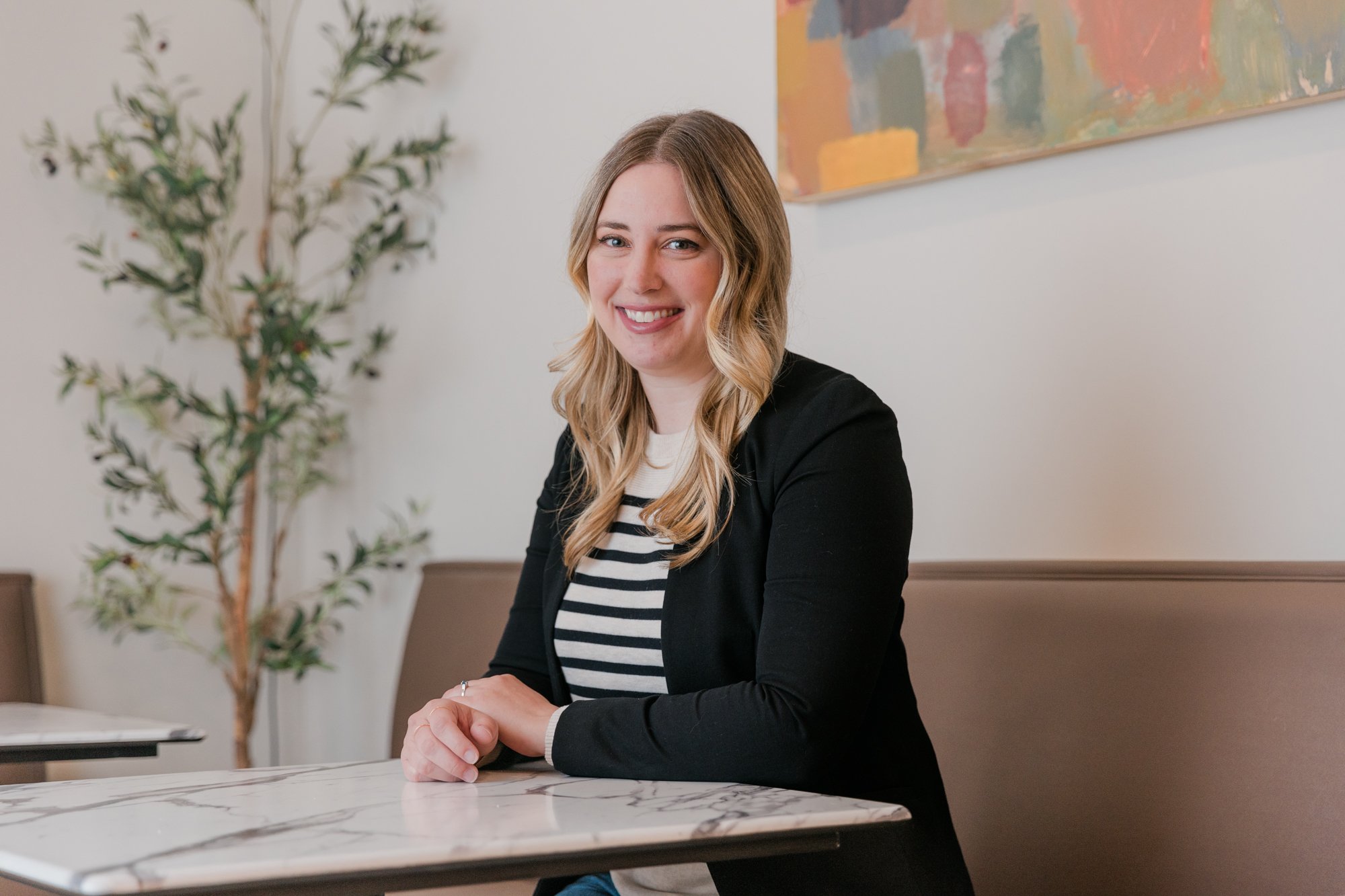 A woman with blonde wavy hair and a striped shirt, sitting at a marble table, smiling at the camera in a modern interior with a plant and abstract painting in the background.