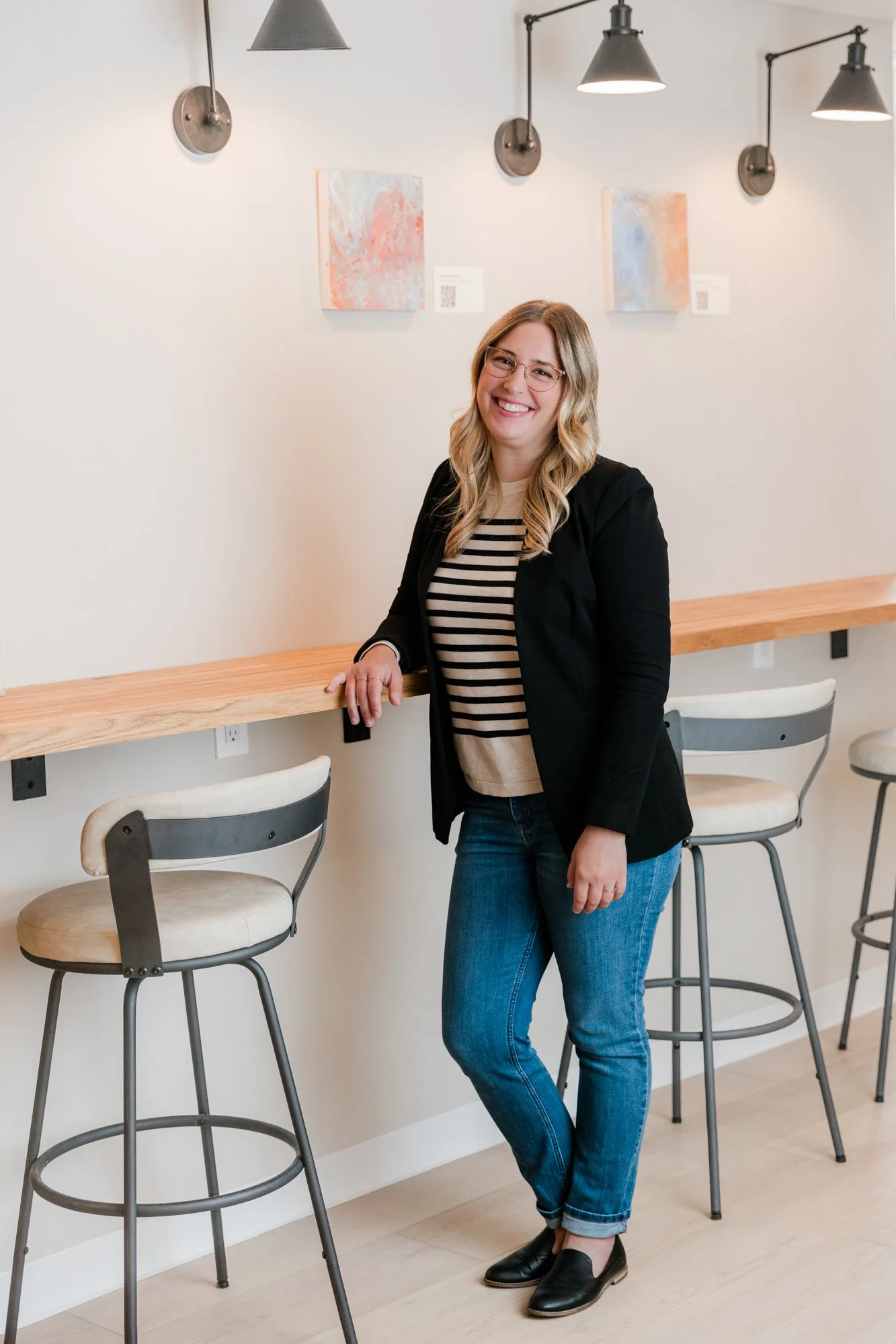 A woman with long blonde wavy hair, glasses, a black blazer, a striped shirt, blue jeans, and black shoes standing inside a cafe or restaurant with high bar stools, a wooden counter, and abstract paintings on the wall.