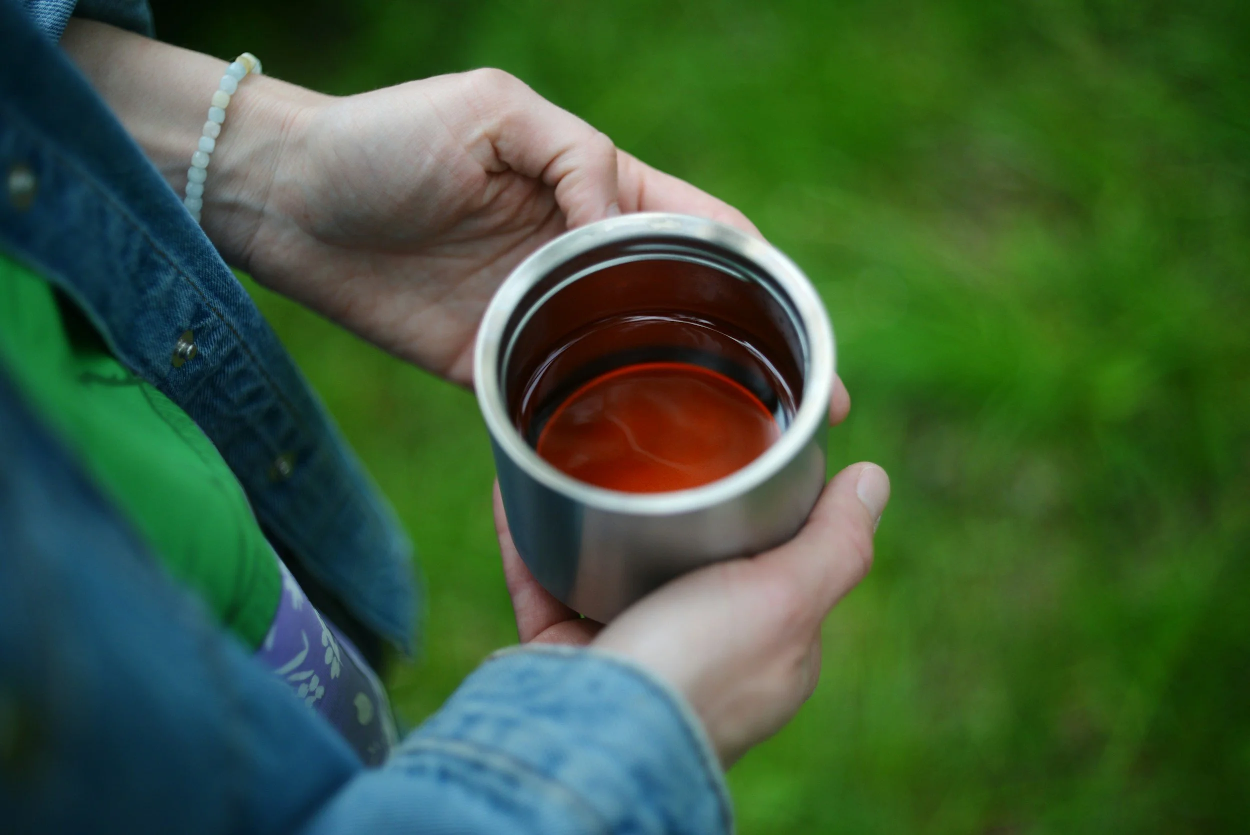 Close-up of a hand holding a silver travel mug with a dark brown beverage, against a background of green grass and a person wearing a denim jacket and a beaded bracelet.