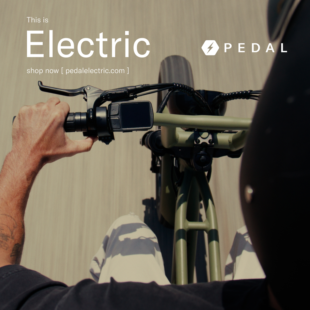 Top-down view of a person riding an electric bike, with one hand on the handlebar, showing the bike's front part and the rider's arm and lap.