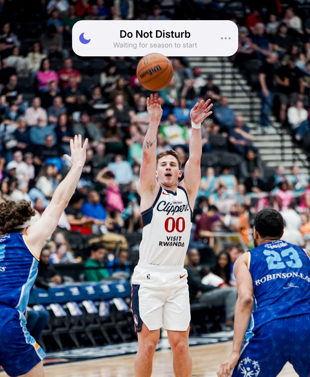 A basketball game with a player in a white jersey shooting the ball while two players in blue jerseys attempt to block.