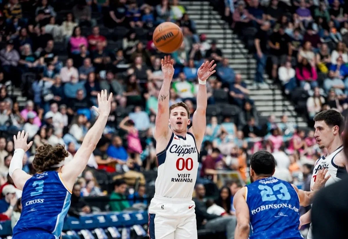 A basketball player in a white jersey with number 00 jumps to shoot the ball while opponents in blue jerseys try to block him during a game in a crowded gymnasium.