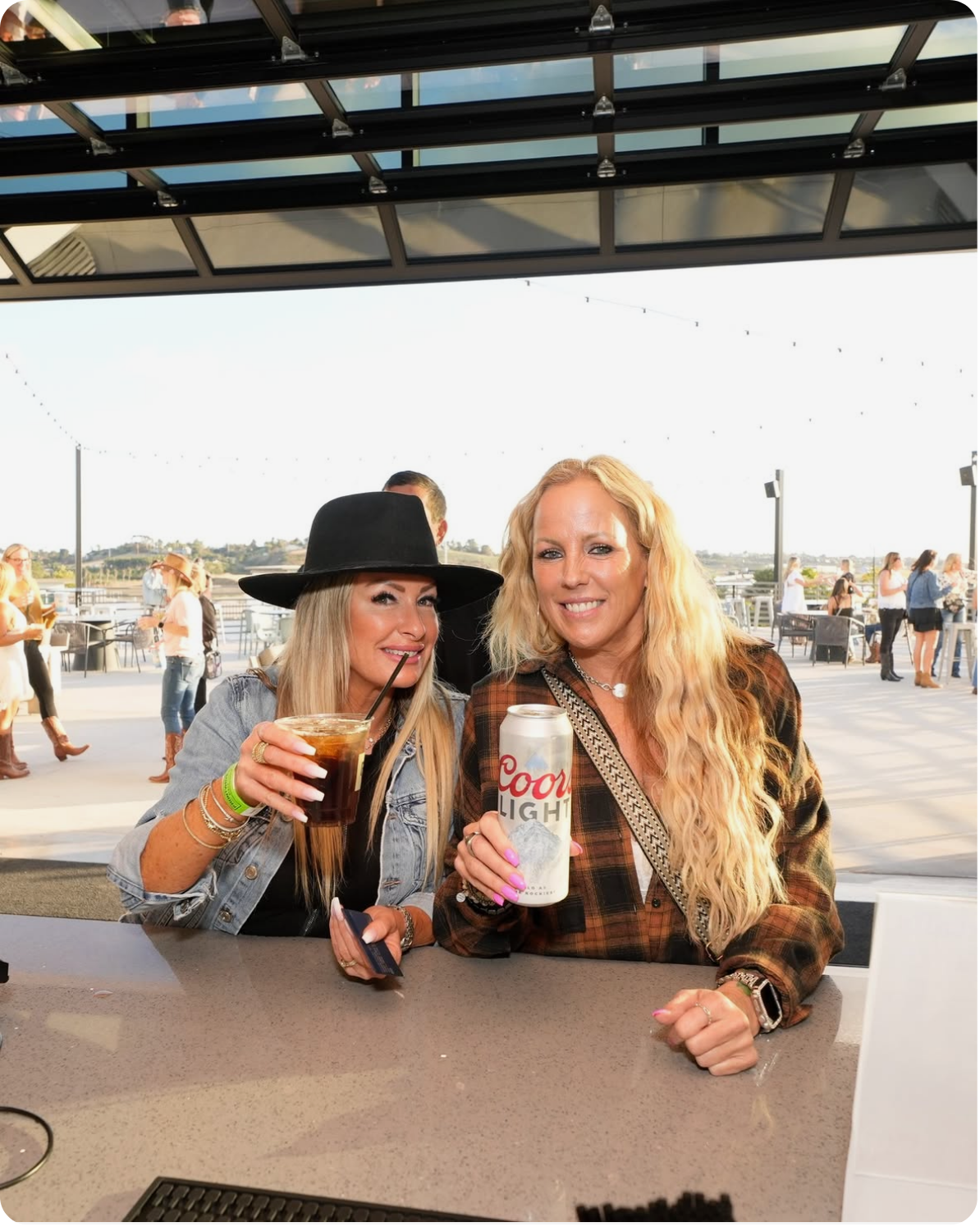 Two women sitting at a bar on a rooftop, smiling and holding drinks, with other people in the background and cityscape views.