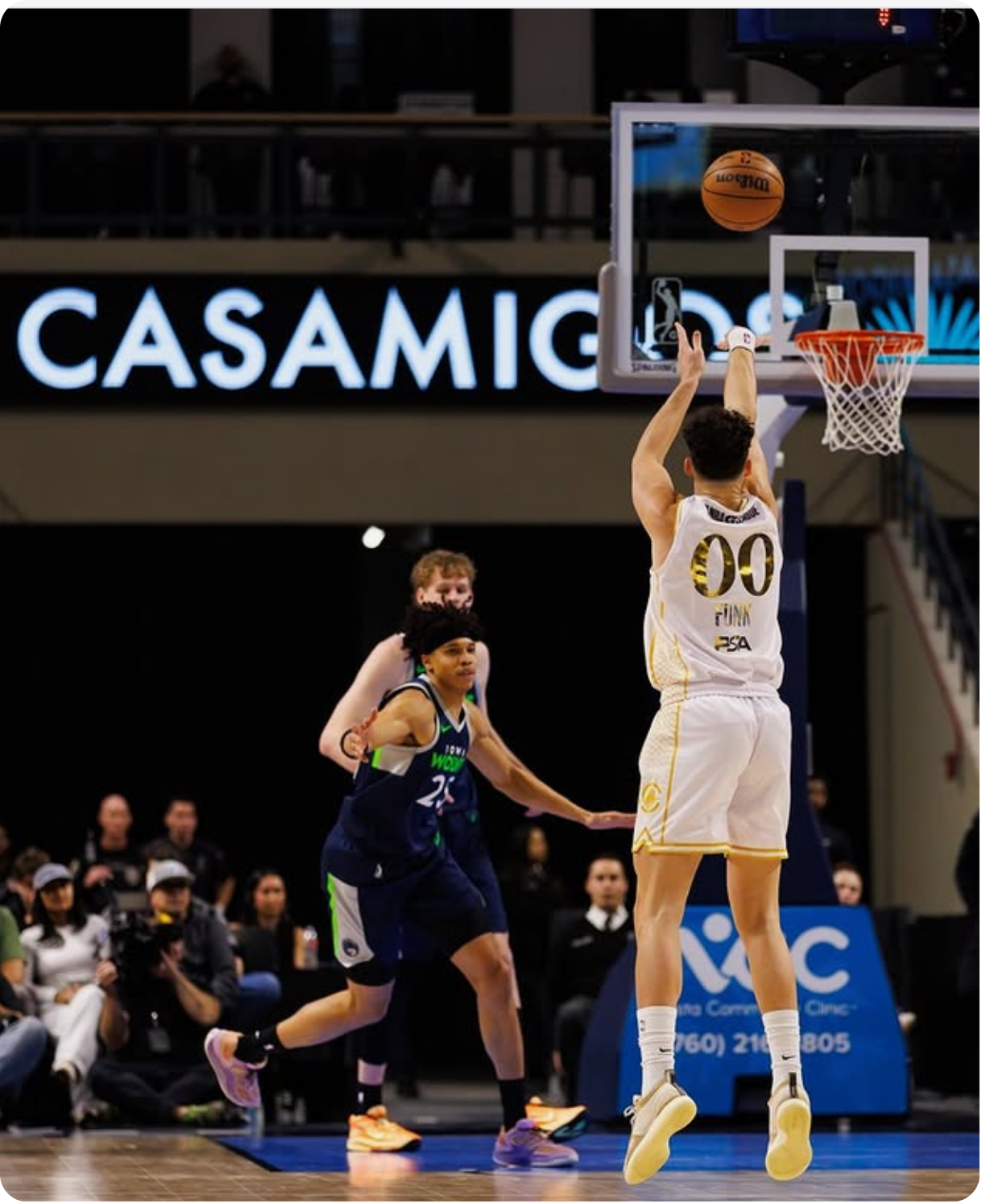 A basketball game in progress with a player shooting the ball towards the hoop and two players attempting to block or defend, with spectators and a blue advertisement banner in the background.