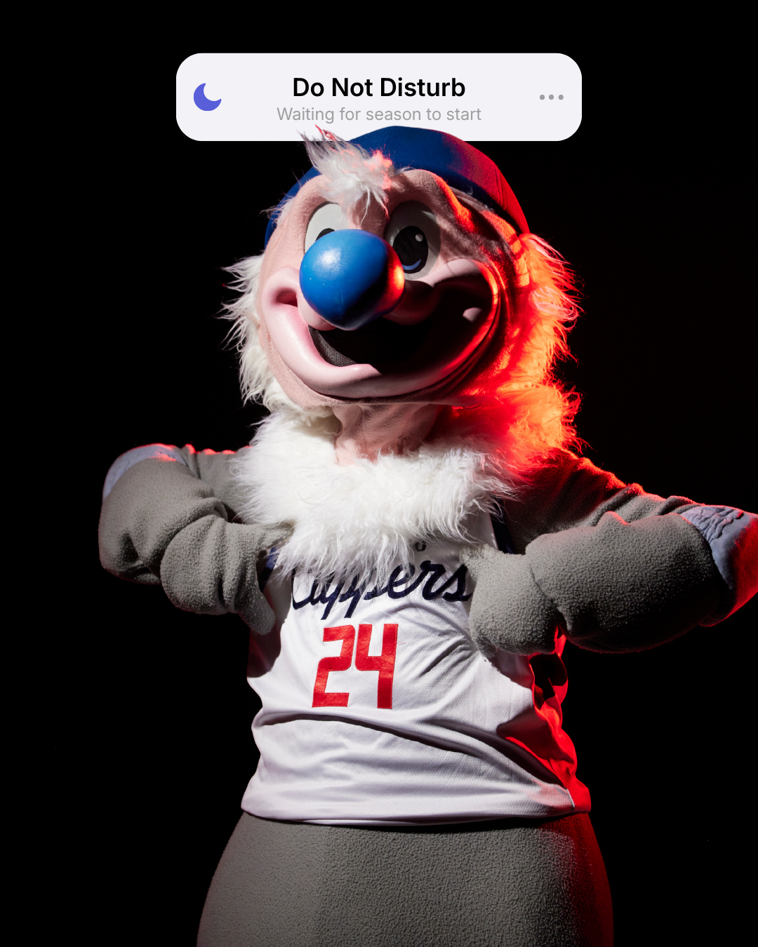 Costumed sports mascot with a large blue nose, white fur, and grey gloves, wearing a white jersey with red and black lettering and the number 24, standing in a spotlight against a black background.