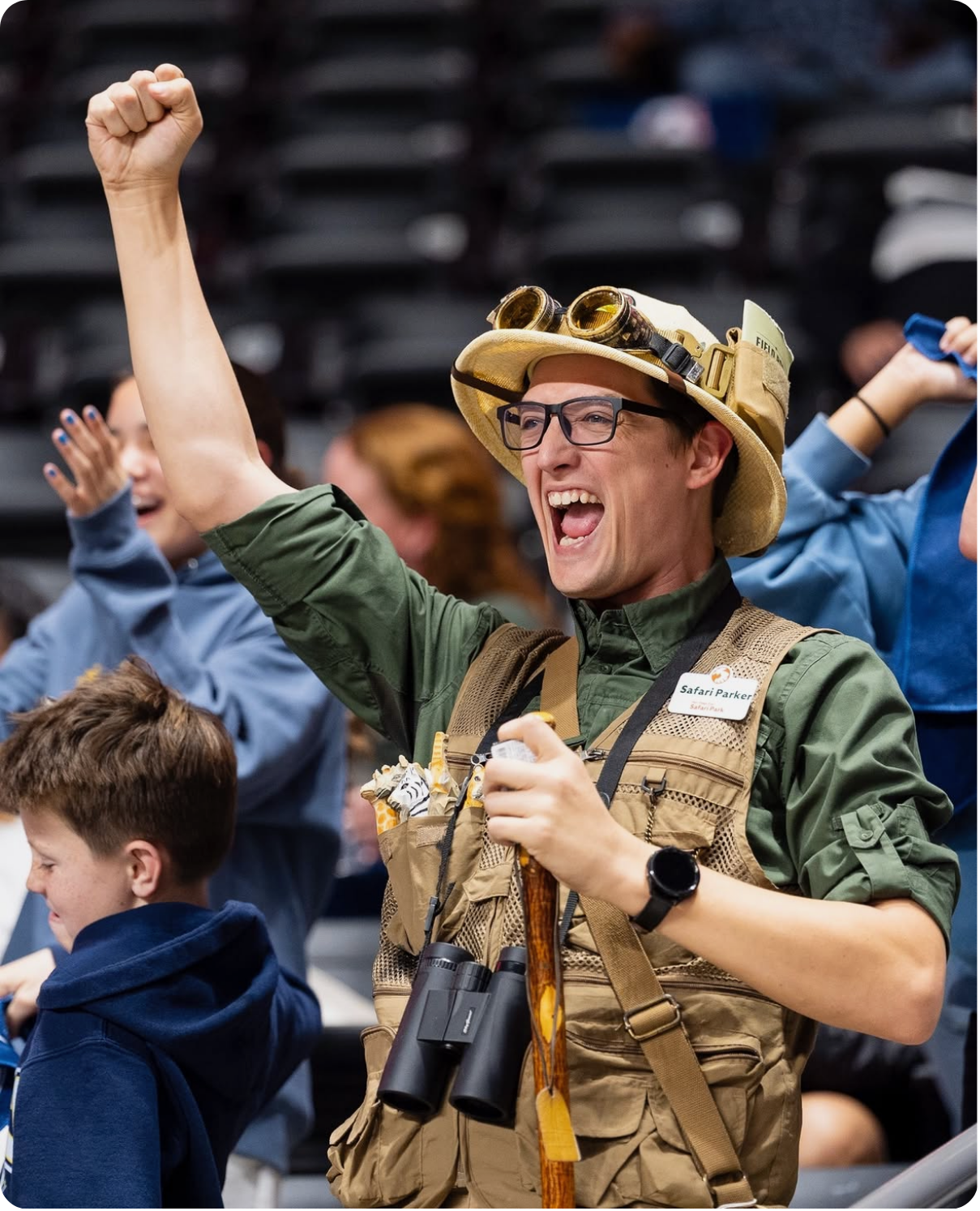 A man wearing glasses, a safari hat with goggles, and a tan vest is raising his fist in excitement while holding binoculars with a name tag that says "Safari Parker". He is smiling and appears to be at an outdoor event or safari-themed gathering.