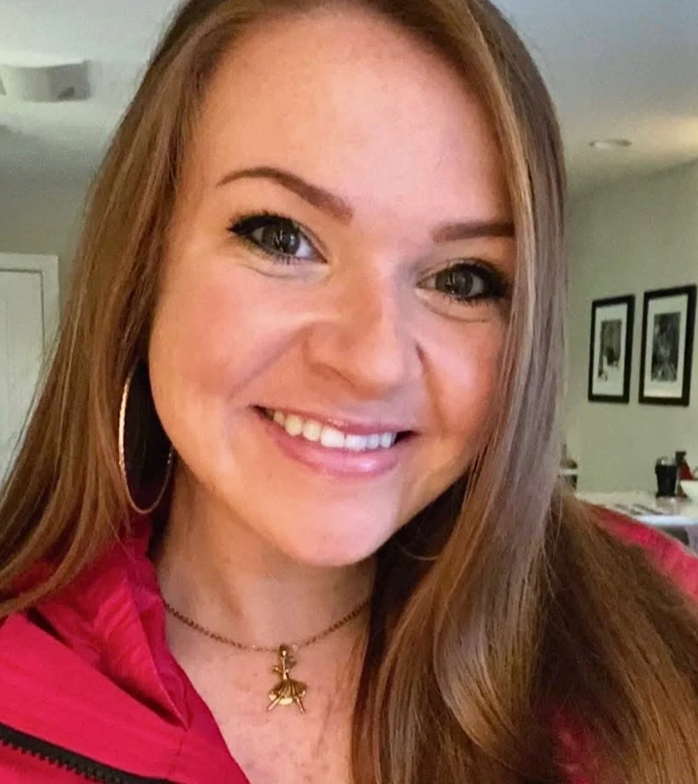 Close-up of a woman with long red hair smiling, wearing hoop earrings and a gold necklace with a bee charm, in a room with framed pictures on the wall.