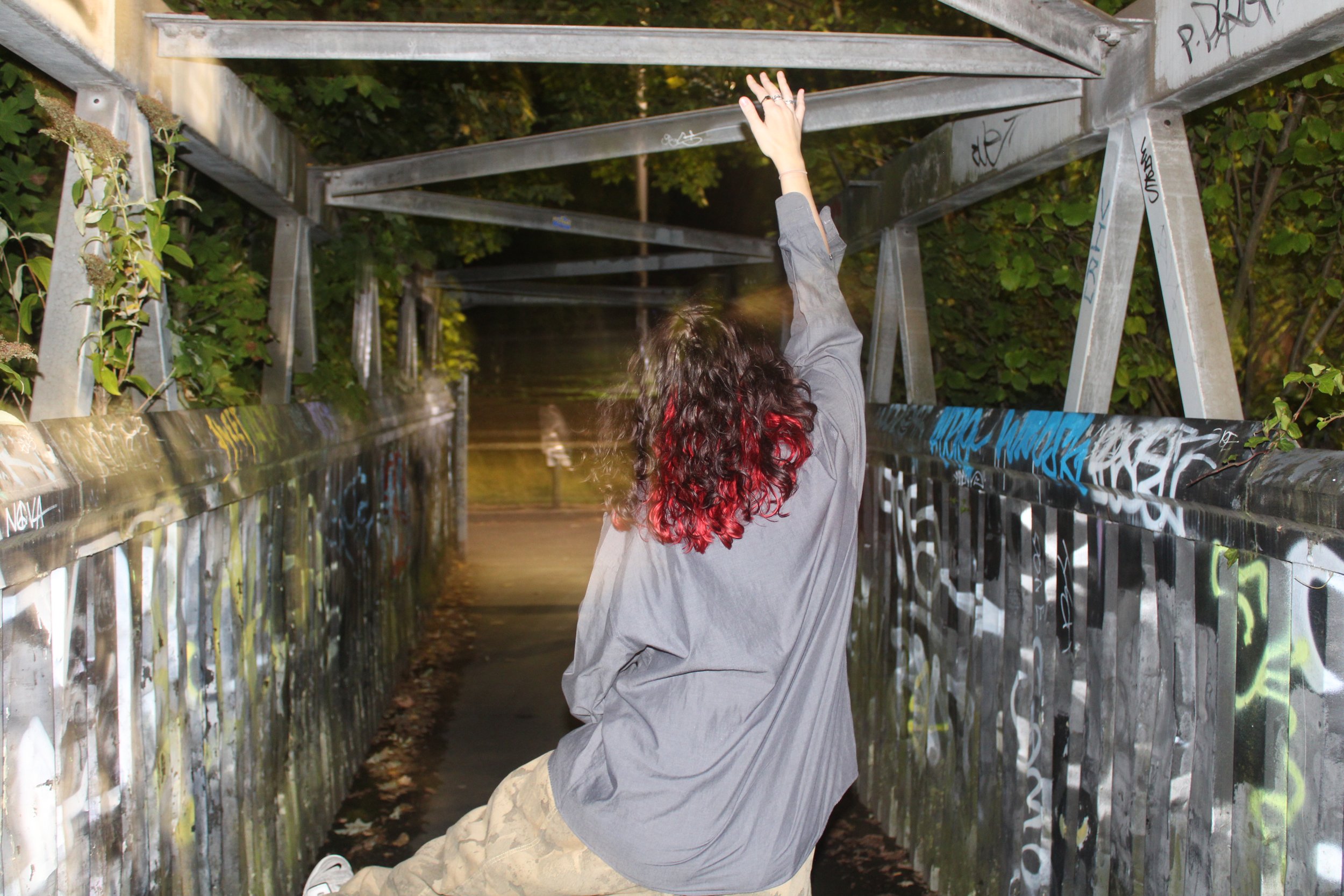 Person with curly hair reaching upward inside a graffiti-covered metal tunnel with greenery in the background.
