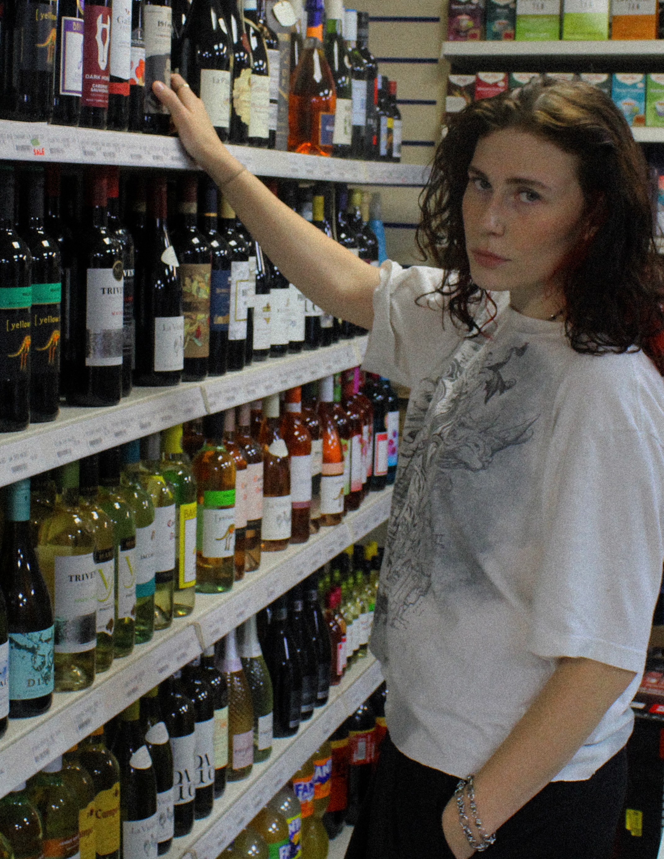 A woman with curly brown hair shopping for wine in a store aisle. She is reaching for a bottle on the top shelf, wearing a white t-shirt with a graphic design, and accessories including bracelets and a watch.