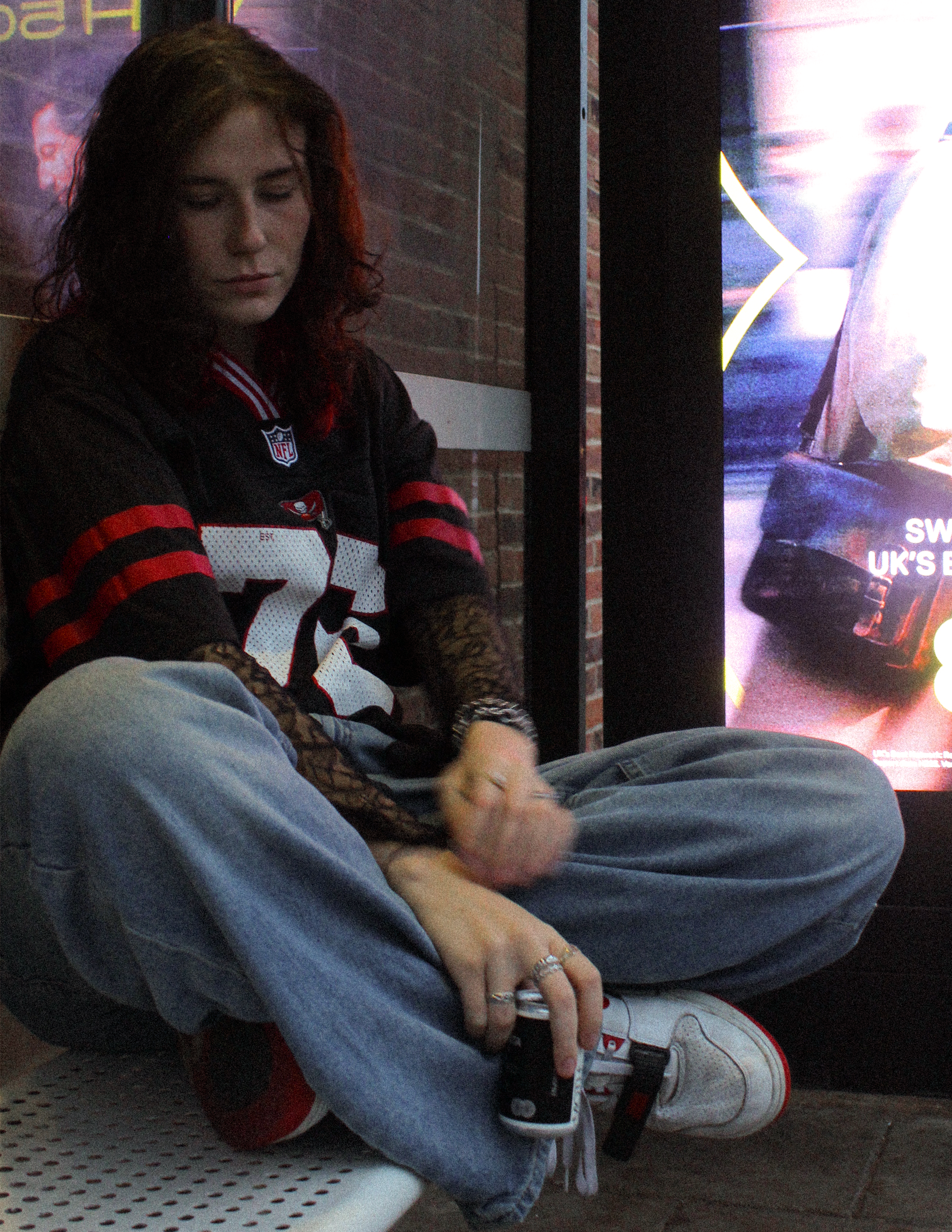 A woman sitting on a white perforated bench, wearing a black and red sports jersey, gray sweatpants, and white sneakers, with her hand resting on her knee.