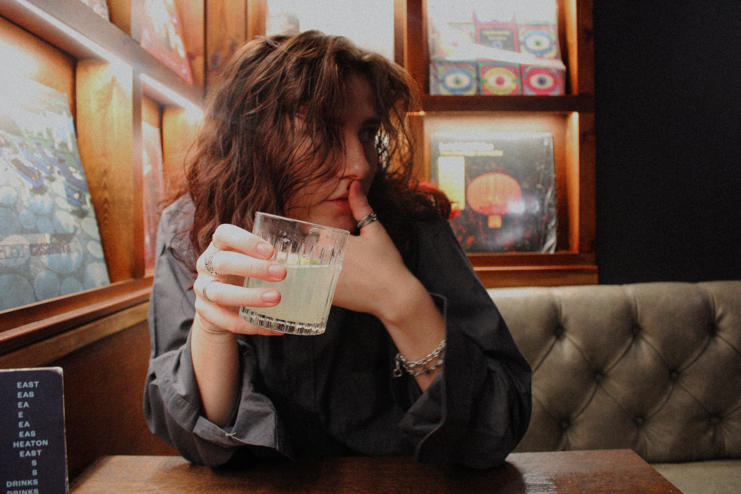 A woman with curly brown hair holding a glass of what appears to be a light-colored beverage, sitting at a wooden booth in a restaurant or bar. She has rings on her fingers and is wearing a dark top. In the background, there are colorful posters and a menu on a small blackboard.