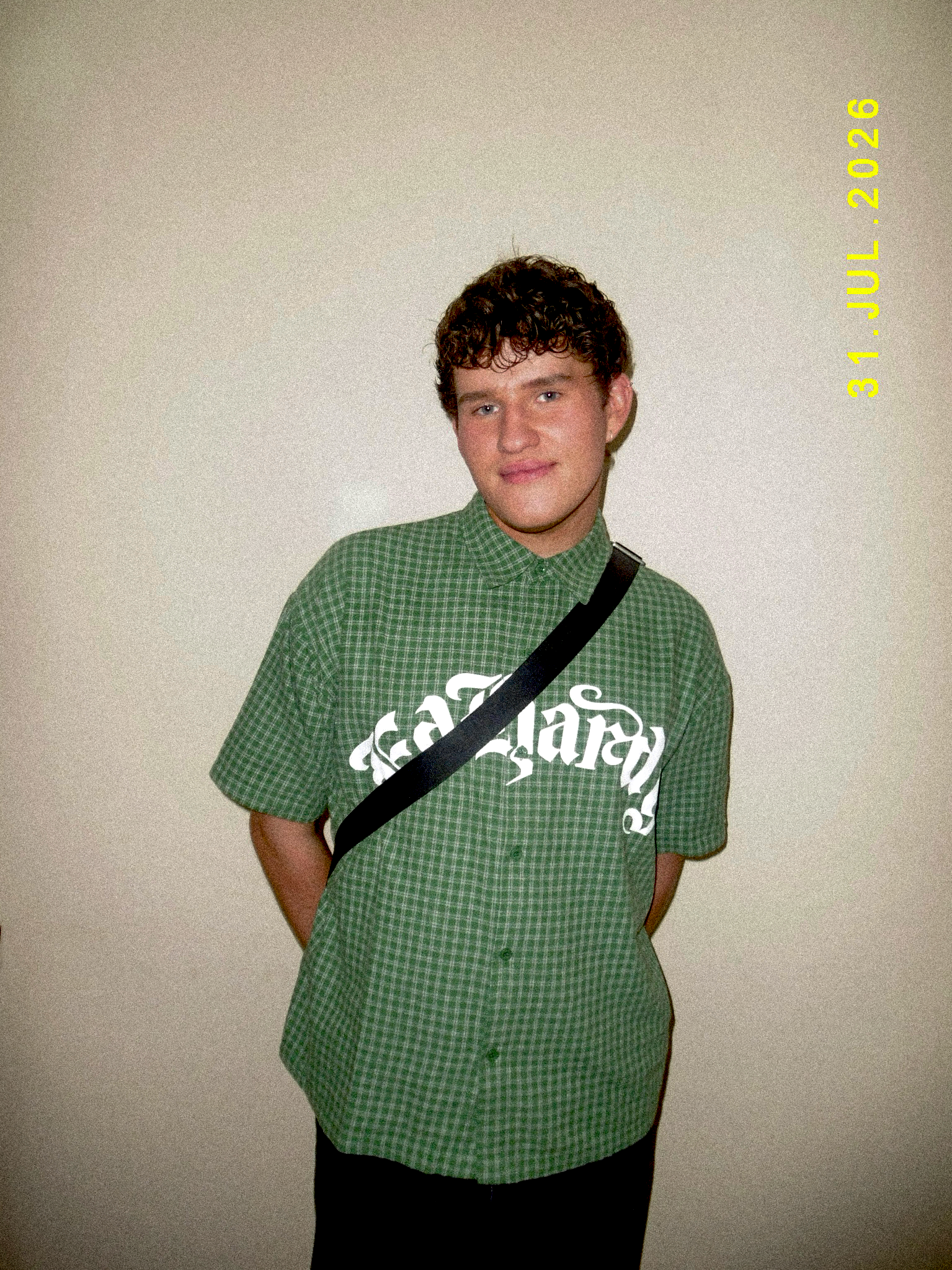 A young man with curly brown hair and light skin, smiling, wearing a green checkered shirt with white lettering and a black strap across his chest, standing against a plain white wall. The date 'July 26, 2020' is visible in yellow text on the right side of the image.