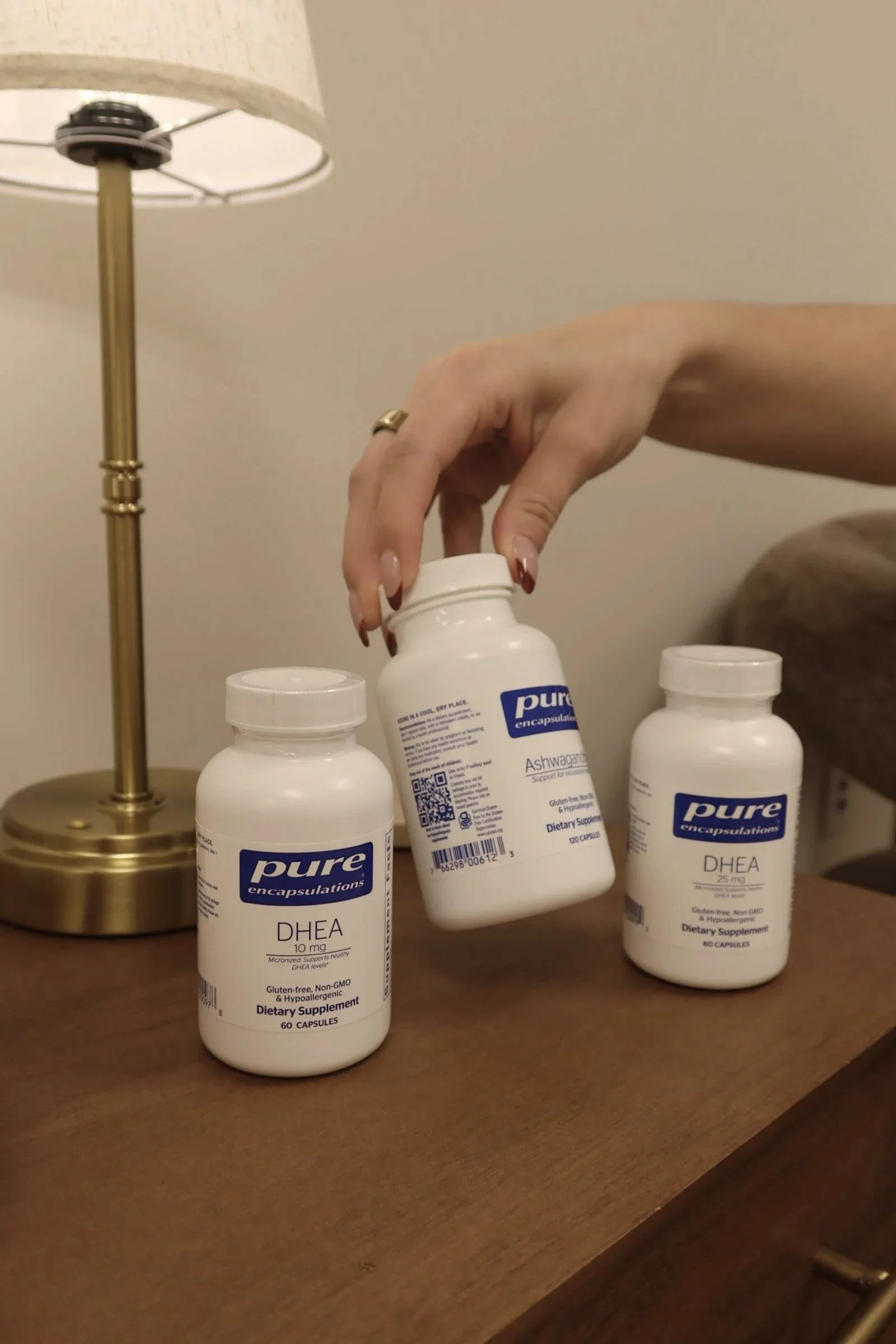 Several bottles of dietary supplements labeled 'Pure Encapsulations DHEA 10 mg' on a wooden table, with a gold-colored lamp in the background.