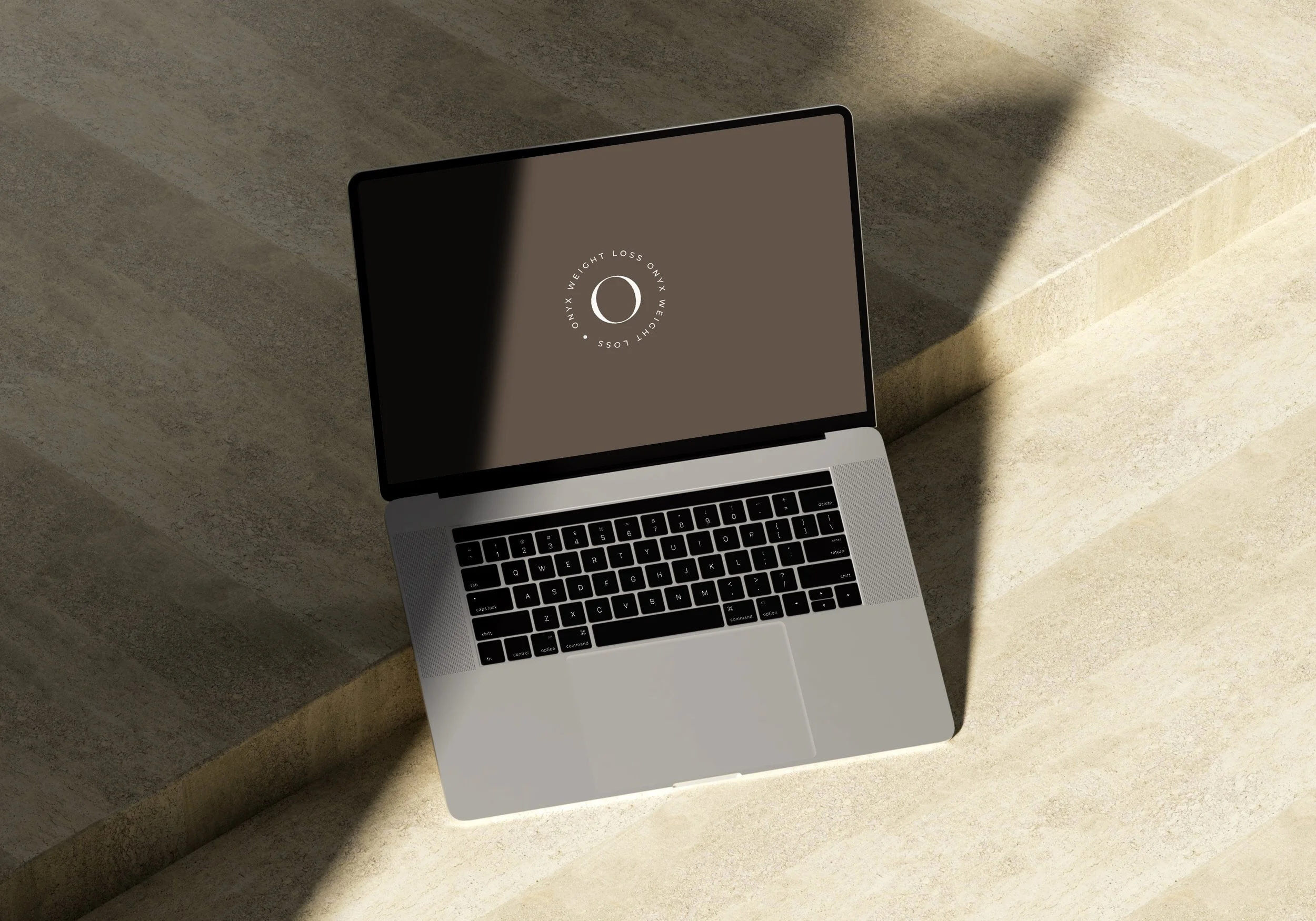 A silver laptop with an open screen displaying a circular logo with text, placed on a beige stone surface. Shadows cast on the surface from light source.