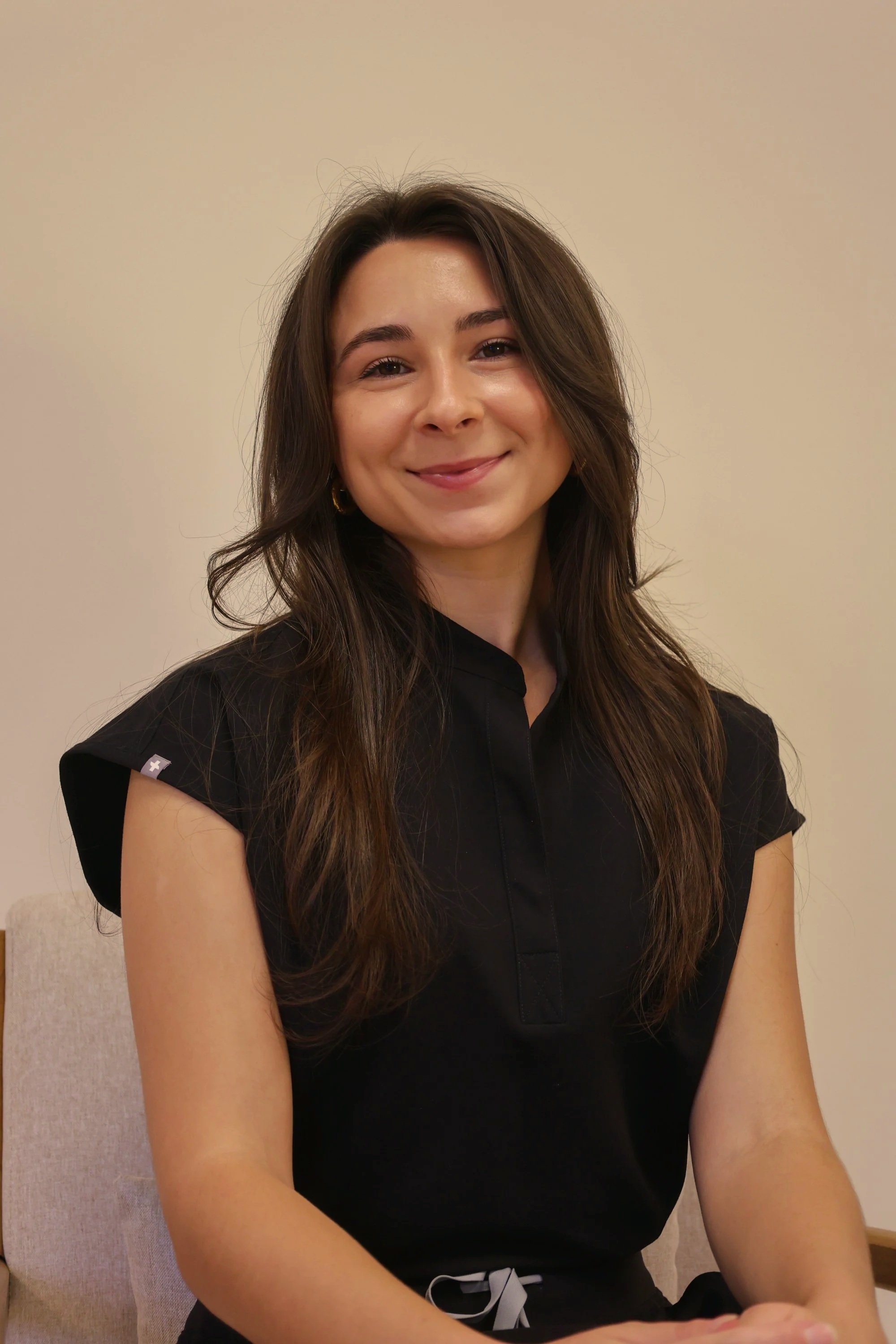 A young woman with long brown hair, smiling and wearing a black short-sleeved top, sitting on a beige chair against a plain off-white wall.