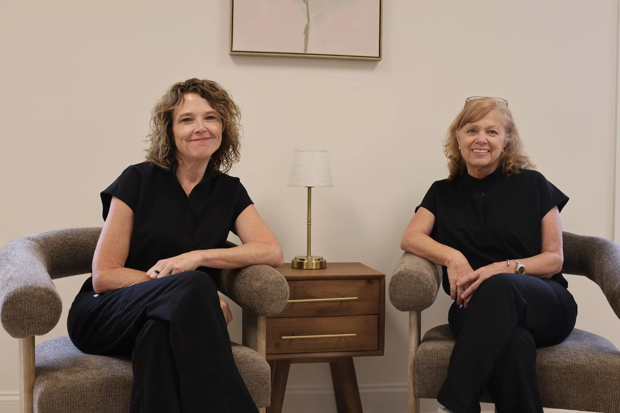 Two women sitting on beige armchairs in a room with beige walls. They are smiling and wearing black tops. A wooden side table with a gold lamp is between them. A framed abstract painting hangs on the wall above them.