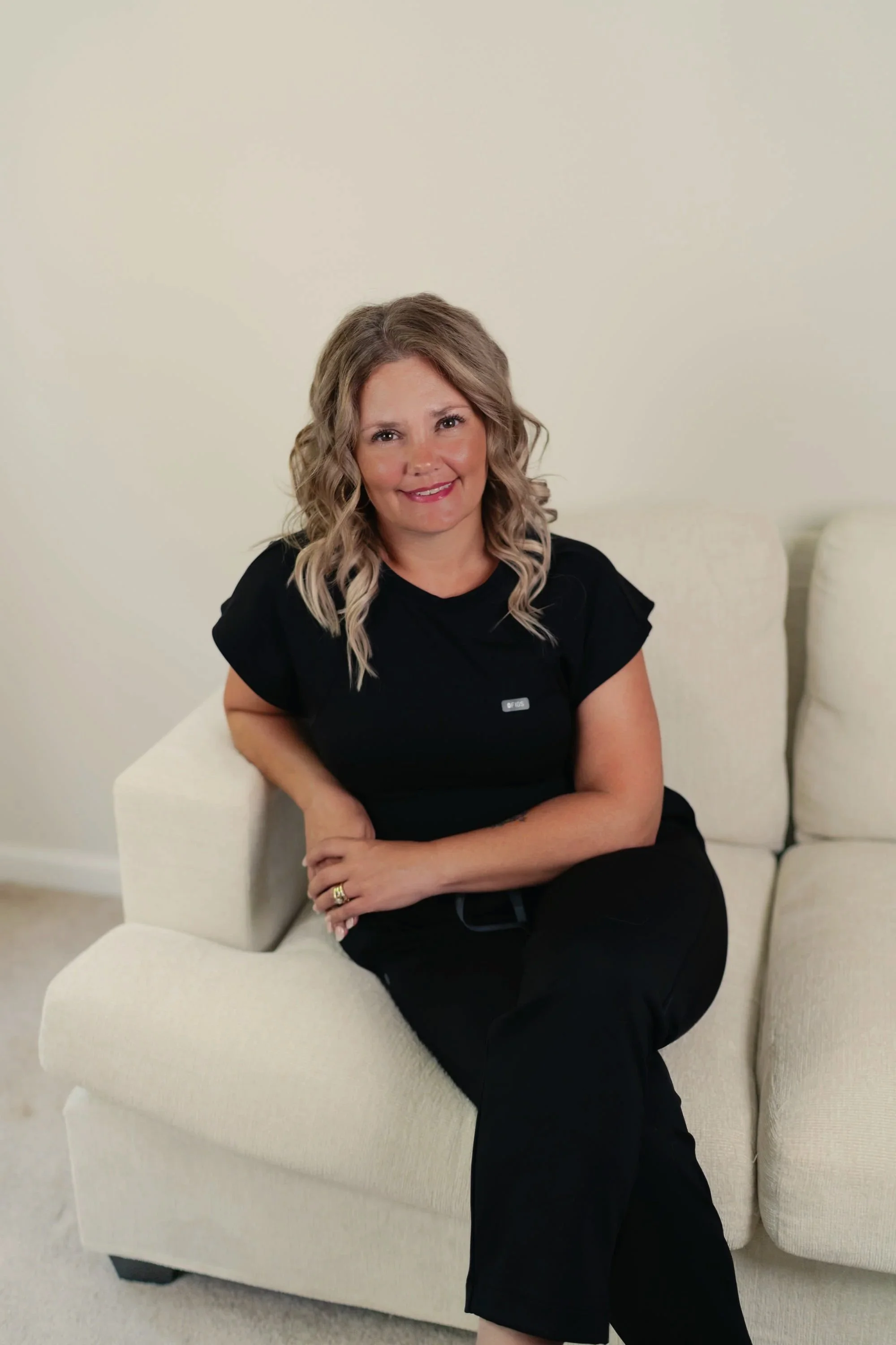 A woman with blonde wavy hair, wearing a black shirt and black pants, sitting on a cream-colored armchair in a room with plain off-white walls, smiling at the camera.