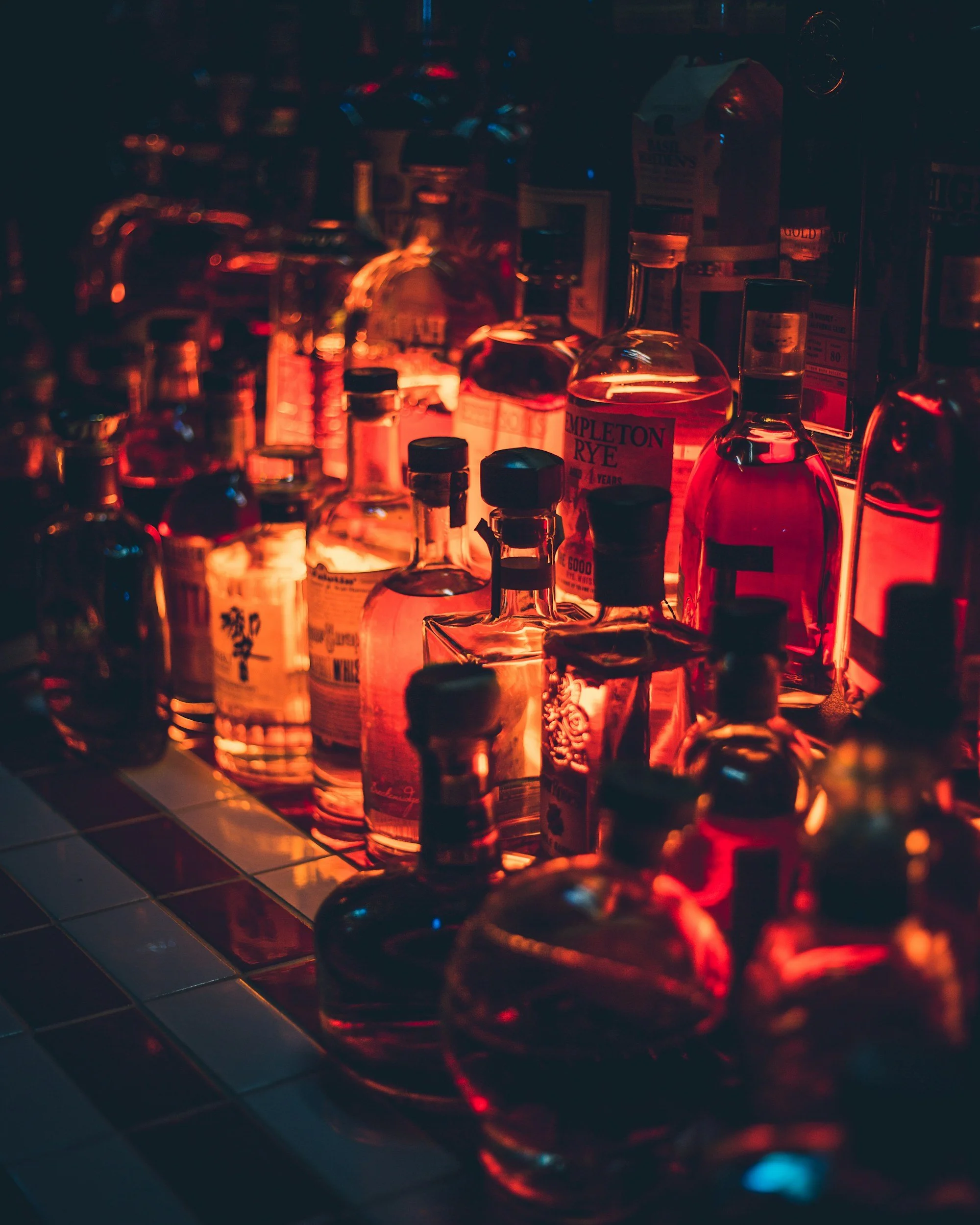 Several bottles of alcohol, including a bottle of Triple Crown Rye, illuminated by red light, on a dark surface, with a tiled floor in the foreground.