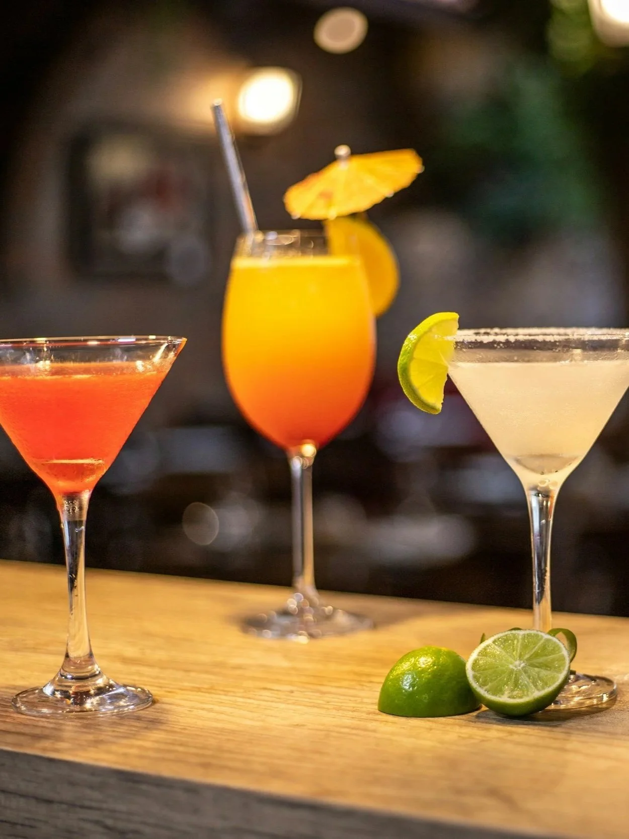 Three colorful cocktails on a wooden bar counter with lime slices and wedges.