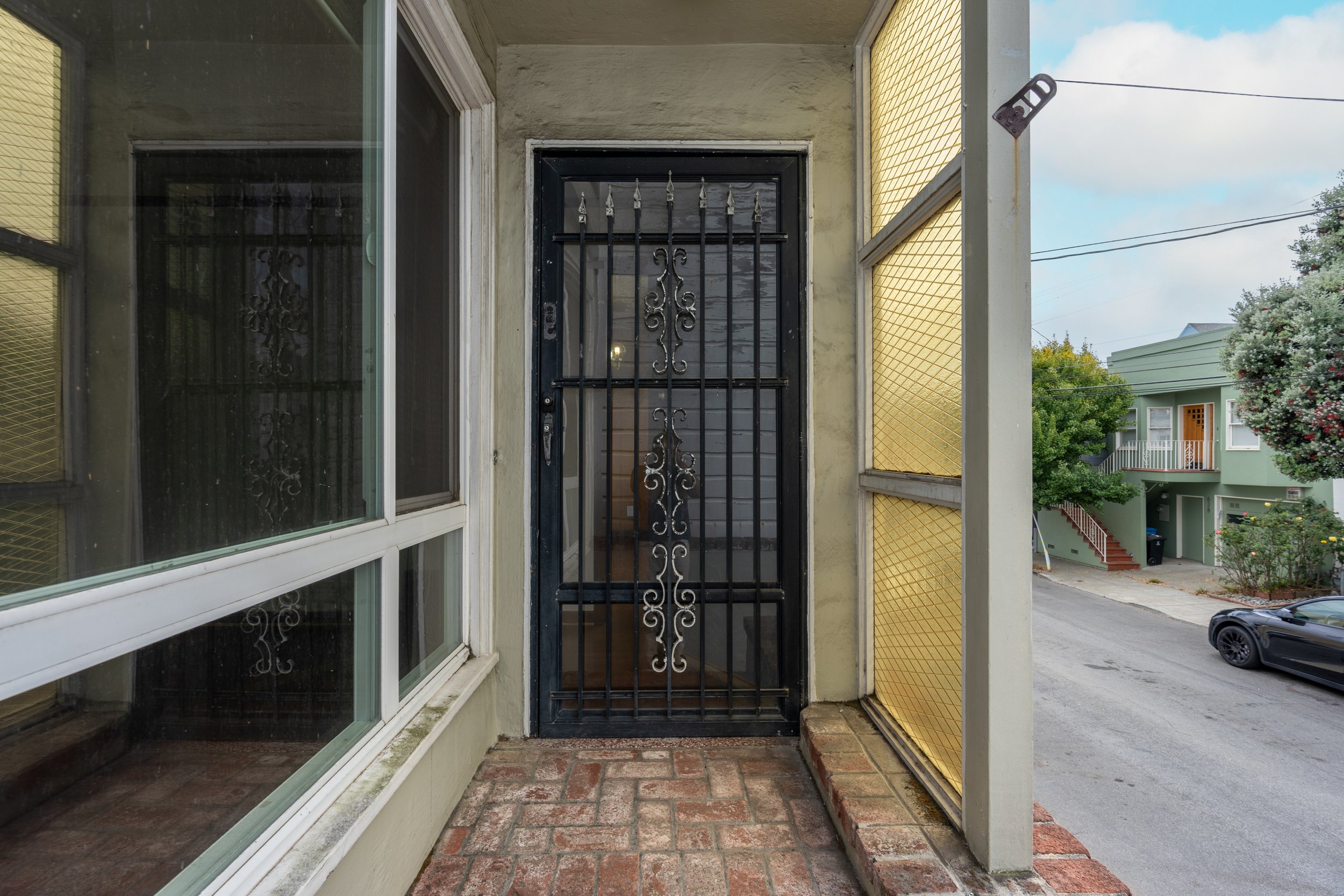 View from a porch showing a black security door with an ornate iron design, alongside large windows and yellow siding, with neighboring houses and a car in the street.