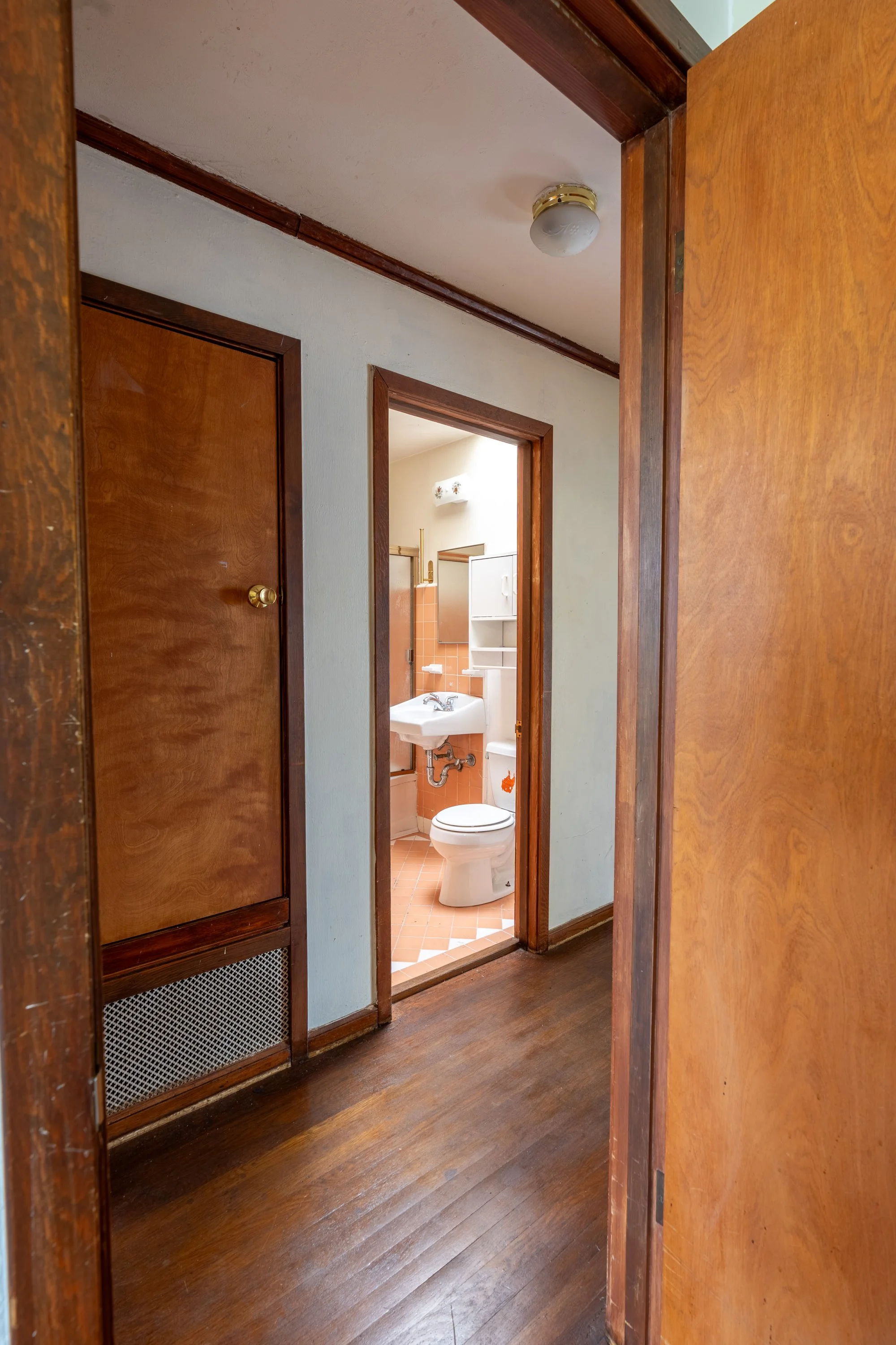A view through a doorway showing a small bathroom with a toilet and a sink with a mirror above it. The bathroom has orange tiles on the floor and a white cabinet. The photo is taken from a room with wooden flooring and brown wooden doors and trim.
