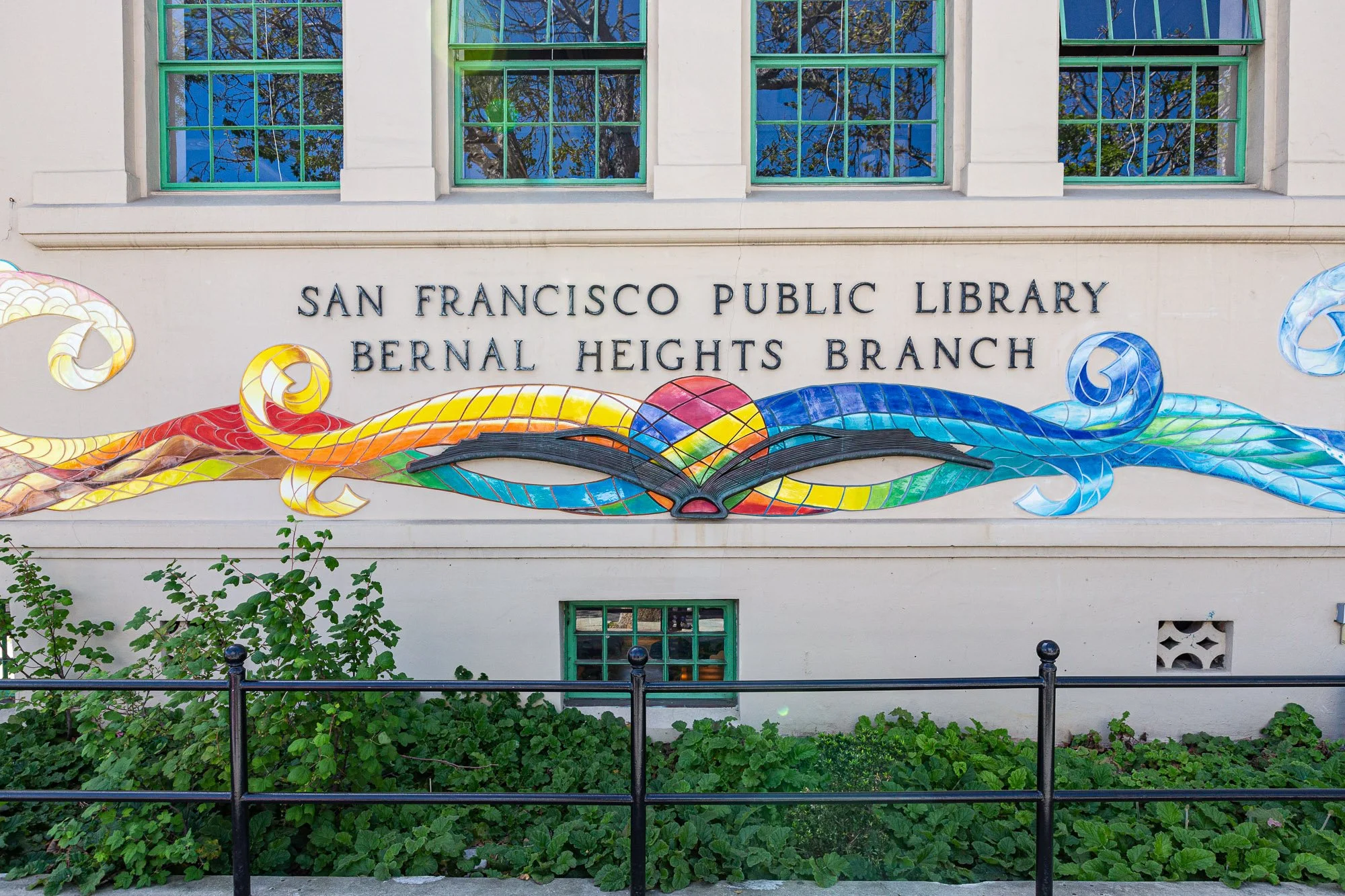 Facade of the San Francisco Public Library Bernal Heights Branch with colorful mosaic art and four windows above, reflected trees visible in the windows.