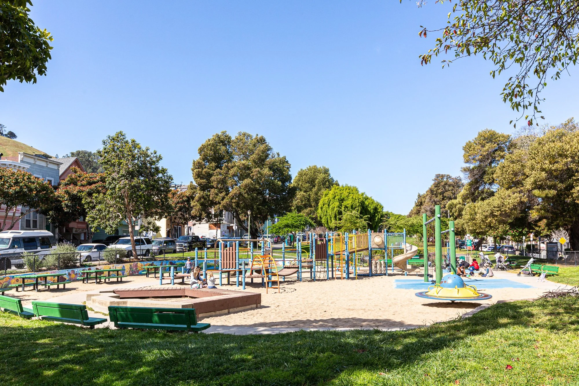 Children playing in a colorful playground with slides, swings, and benches, surrounded by trees and residential buildings in a park during daytime.