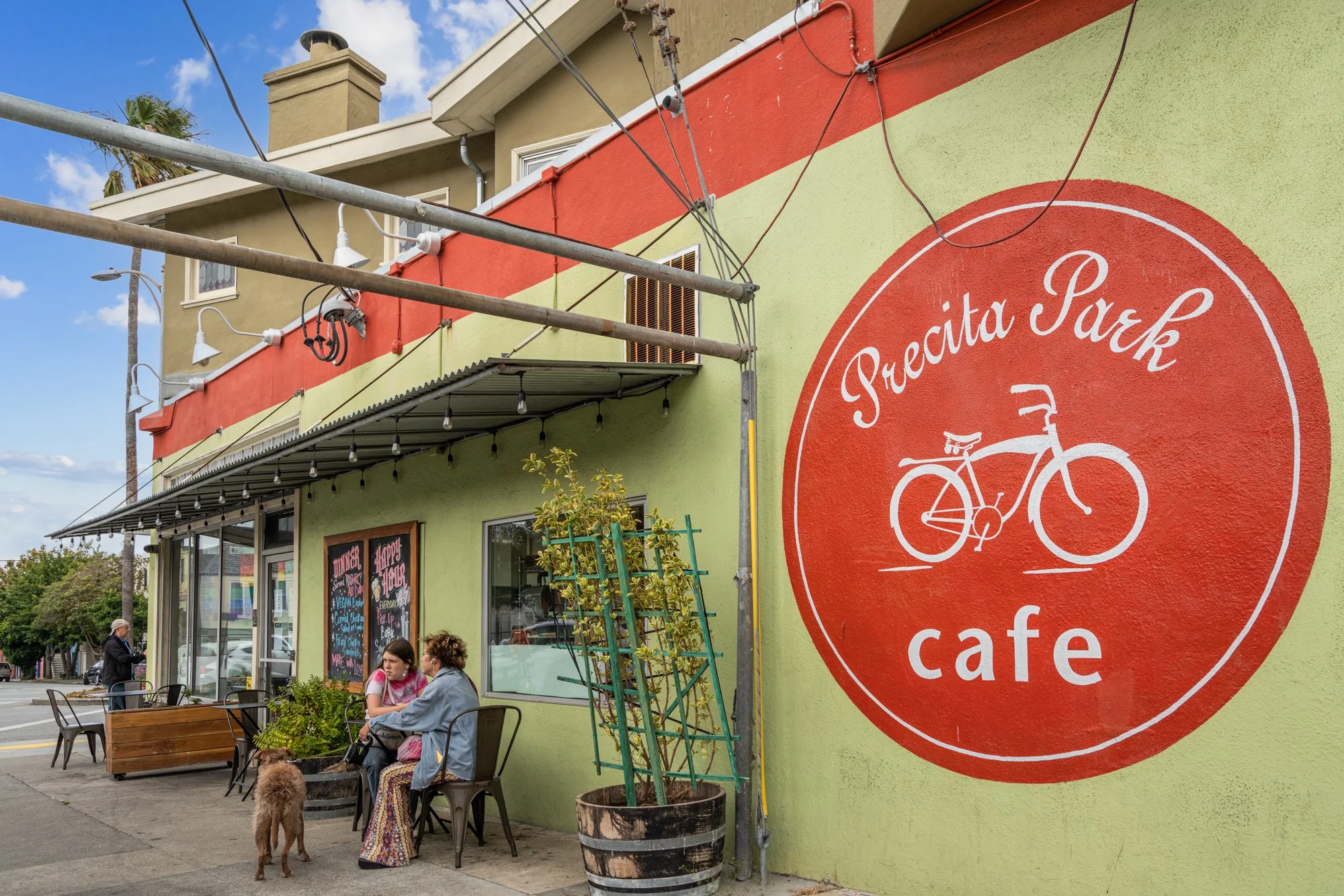 People sitting outside a colorful cafe with a large sign that reads "Proeta Park" and a bicycle graphic. There are tables, chairs, plants, and string lights outside, with a few pedestrians and a dog nearby. The building has a green and red exterior.