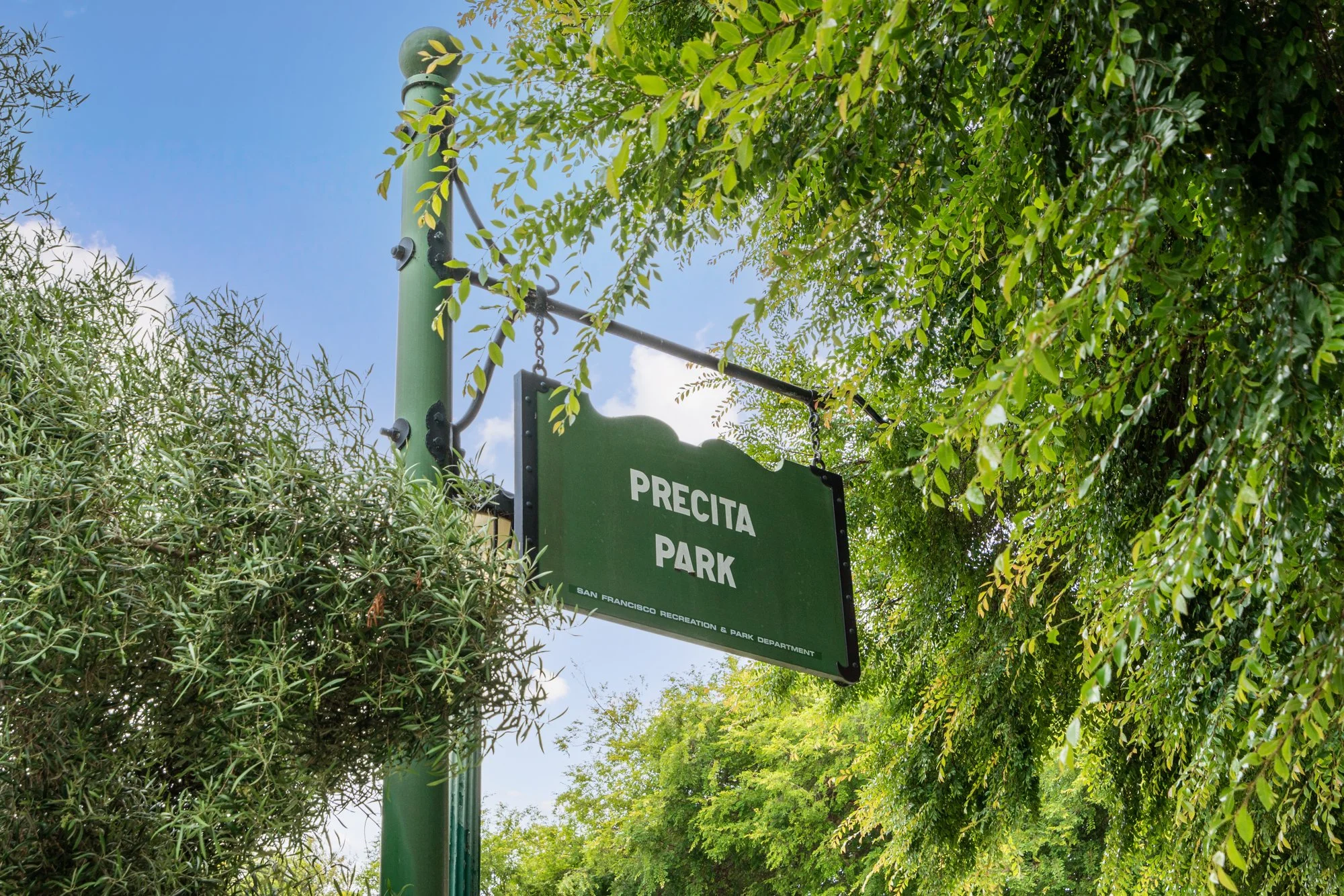 Green park sign reading 'PRECITA PARK' surrounded by lush green trees and bushes. Clear blue sky in the background.