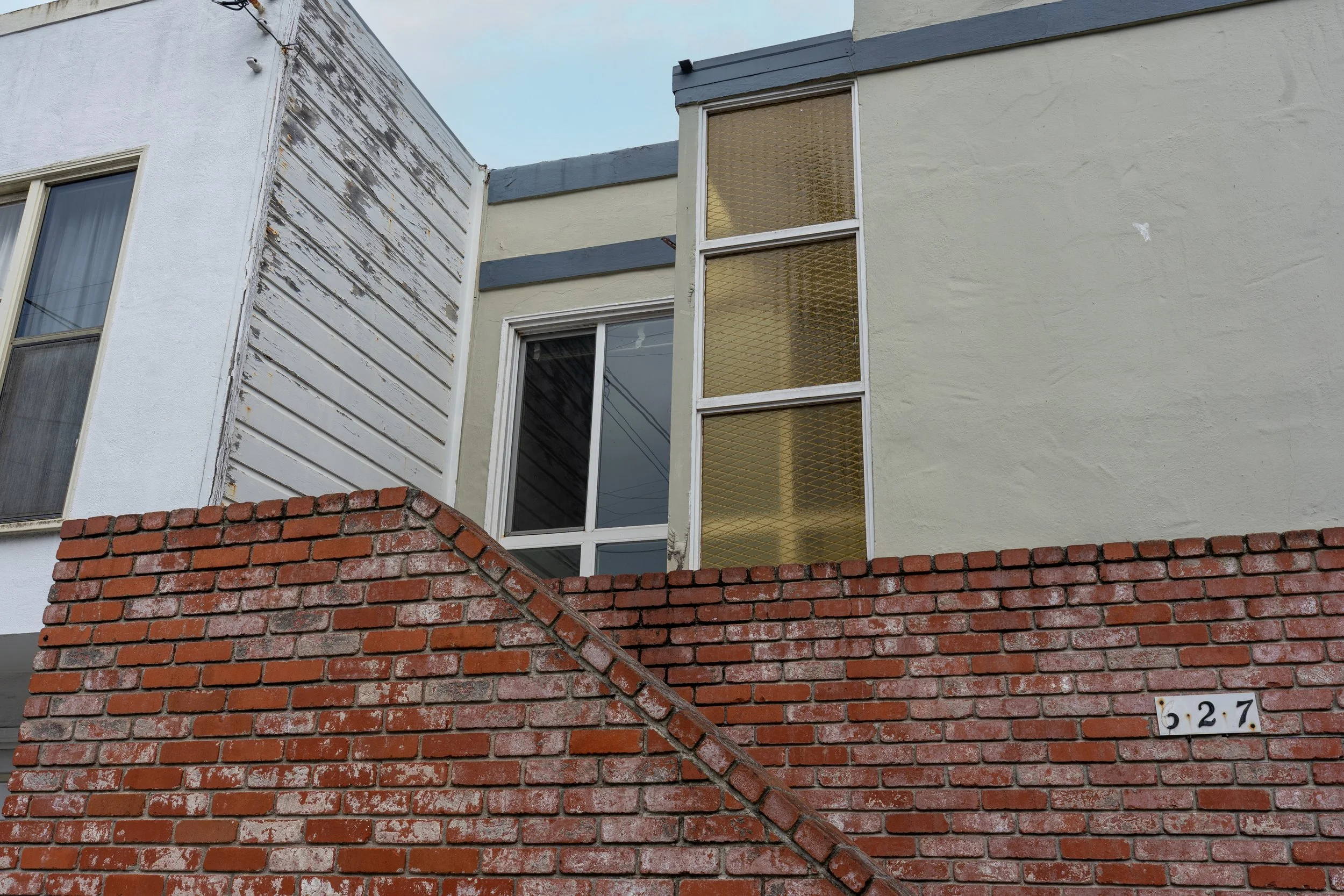 A building with a brick wall in the foreground and upper levels showing white and beige sections with a large window and some weathered wooden siding.