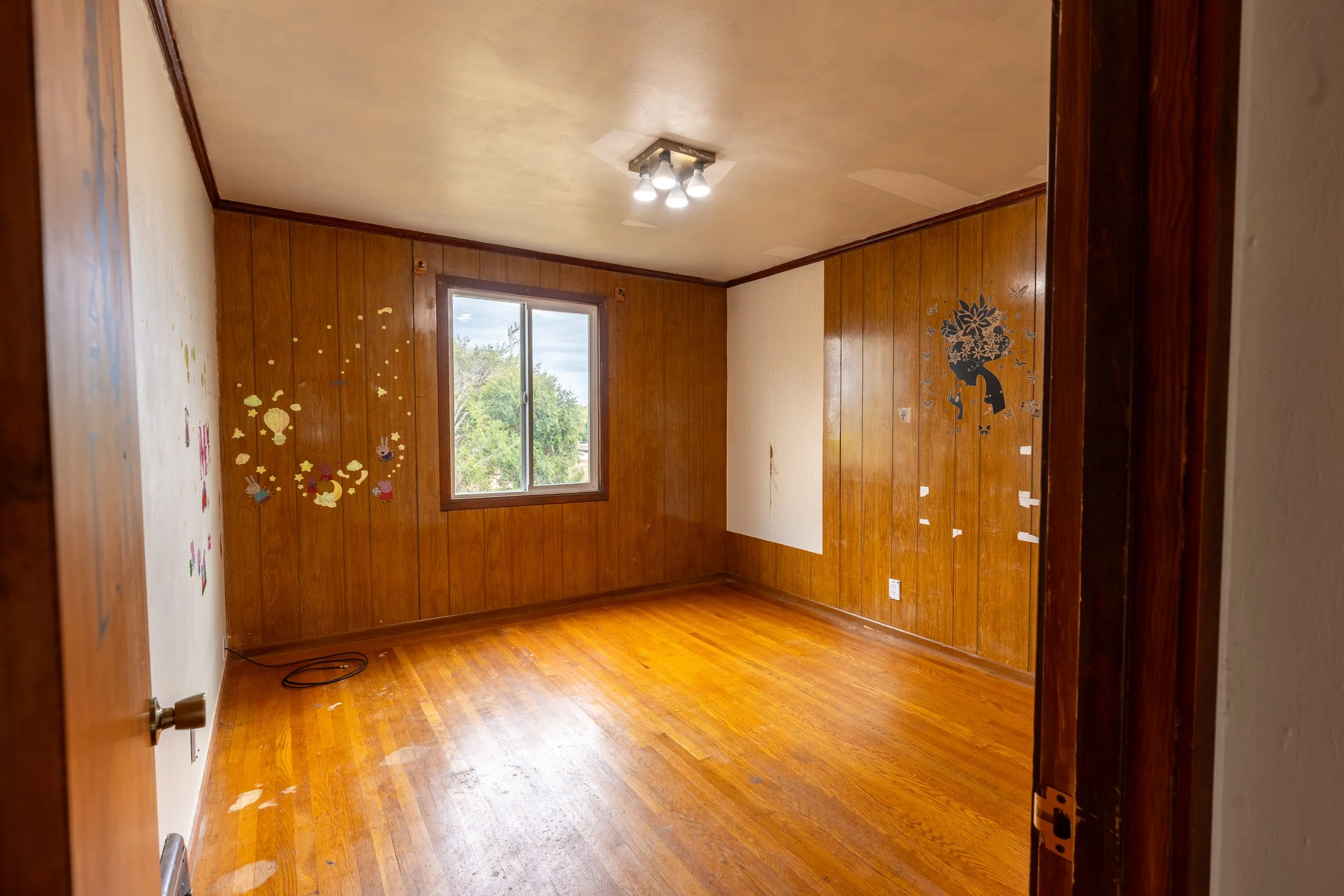 A small, empty room with wooden walls and flooring, a window showing trees outside, a ceiling light fixture with four bulbs, and air vents on the ceiling. There are wall decals of a girl with flowers on her head on one wall, and a partially torn pape