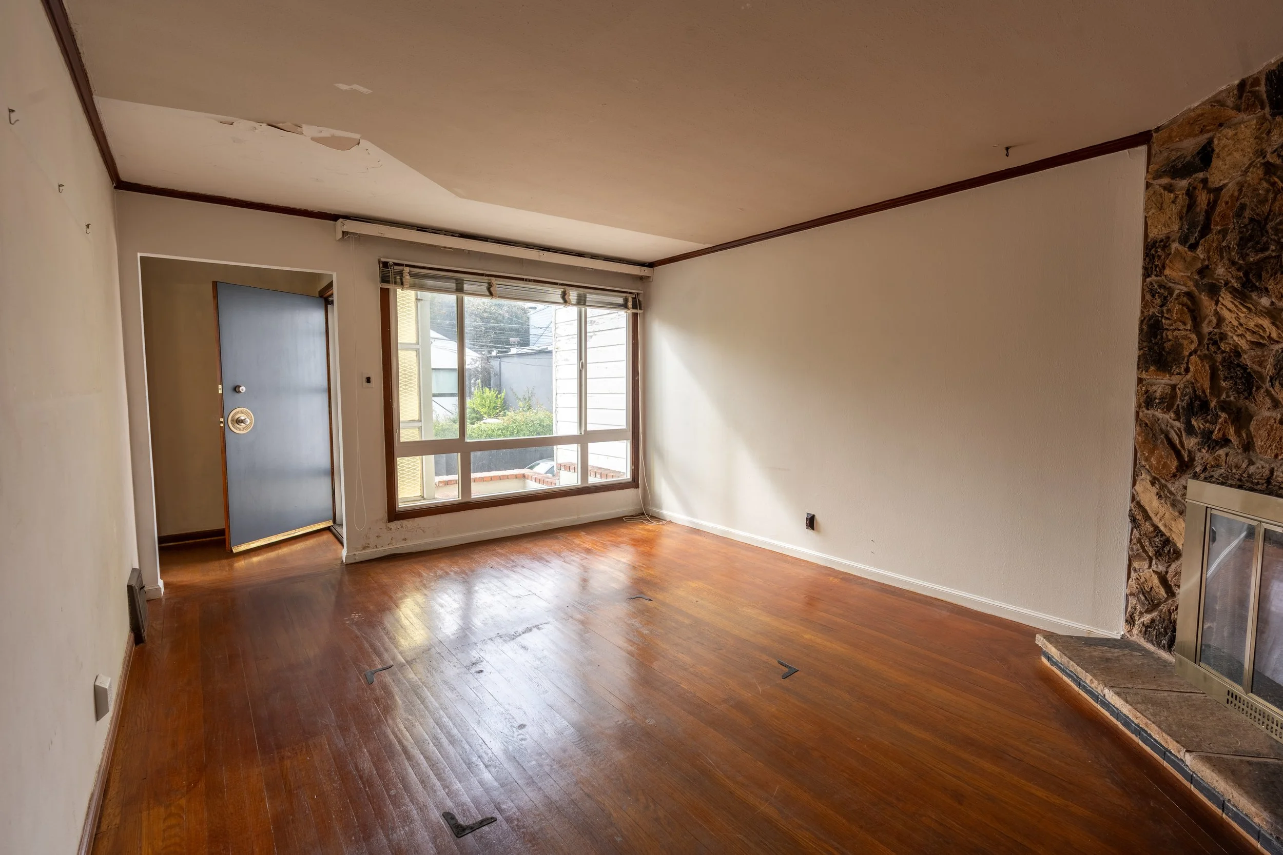 Empty living room with hardwood floors, a large window, a stone fireplace, and a damaged ceiling with peeling paint.