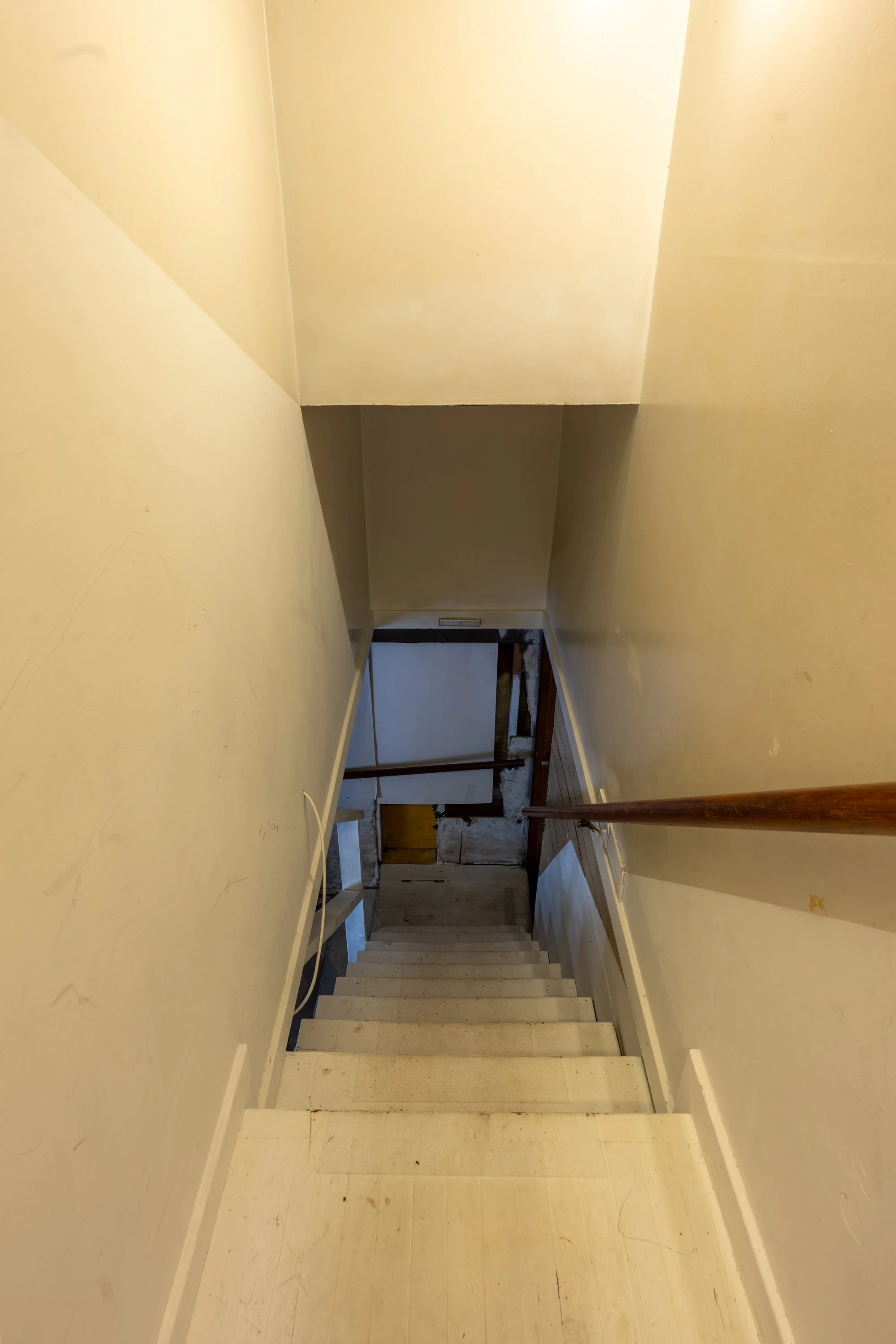 A stairway with a handrail on the right, leading down to a door with a wooden frame. The walls are painted yellow and show signs of wear, with some marks and discoloration.
