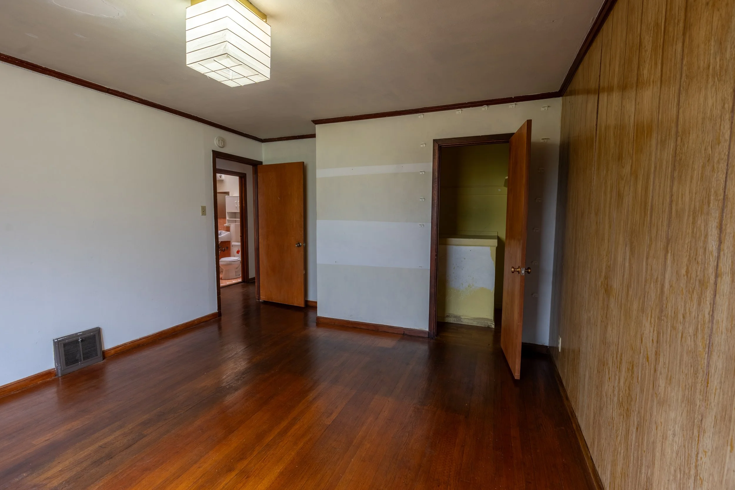 Empty room with hardwood floors, white walls, a wood door leading to a bathroom, and a vent on the floor.
