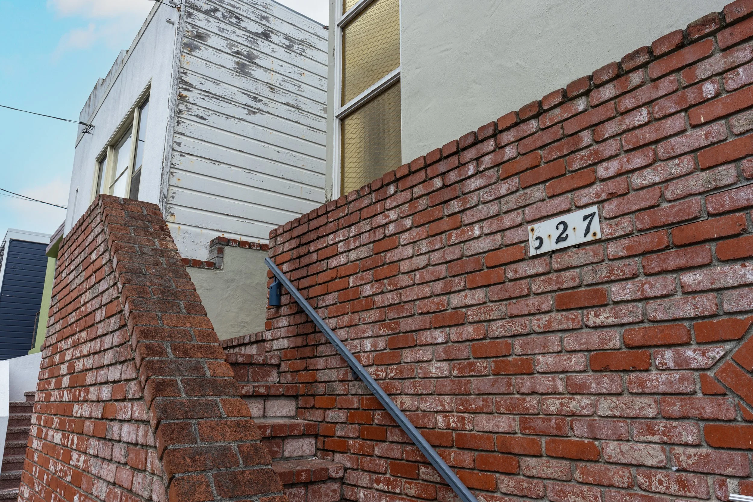 Close-up of a brick staircase with a black metal handrail, leading up to a house with two distinct exterior wall textures: weathered white wood siding and smooth beige stucco, and a house number plaque displaying '627' on the brick wall.