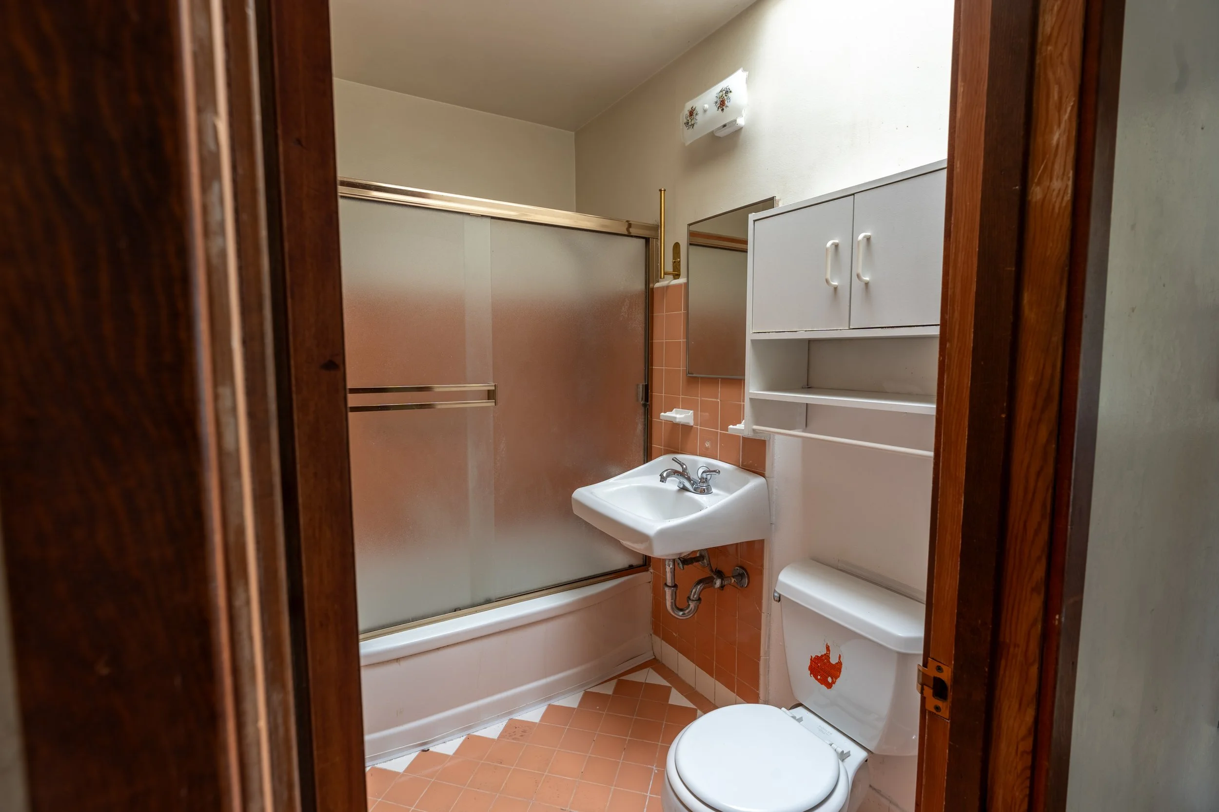 Small bathroom with a shower stall, a white sink, and a toilet. There are white cabinets above the toilet, and a mirror next to the shower. The floor has orange and white tiles, and the walls are painted light green. The photo is taken through a wood