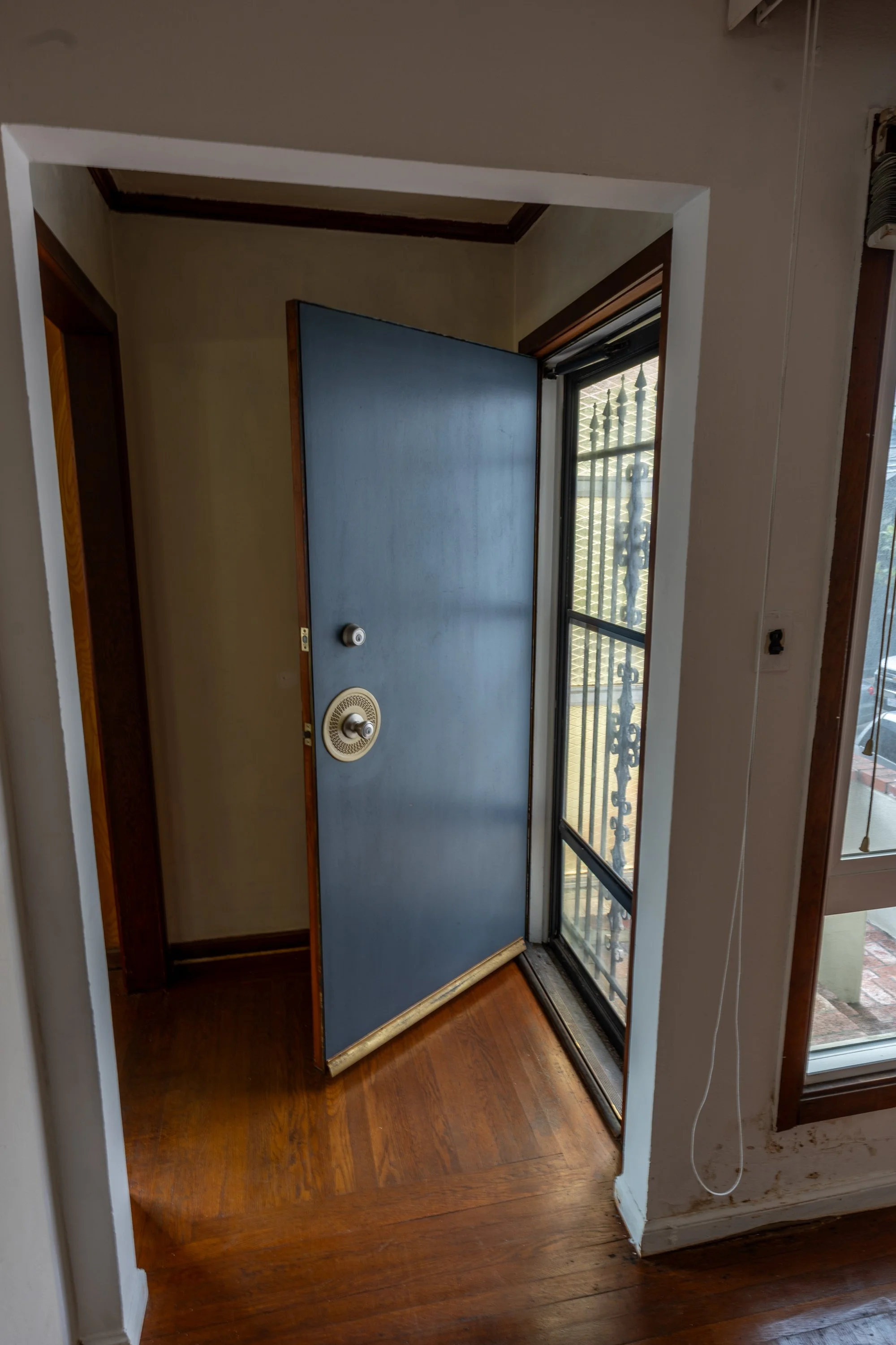 Open front door with a peephole and a screen door, leading to a small hallway with wooden floors and a window with bars outside.
